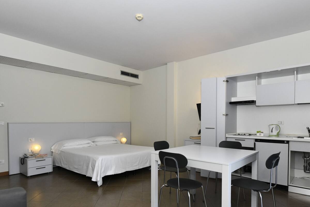 Una camera d'albergo moderna con un letto, comodini, lampada, scrivania, sedie e angolo cottura.