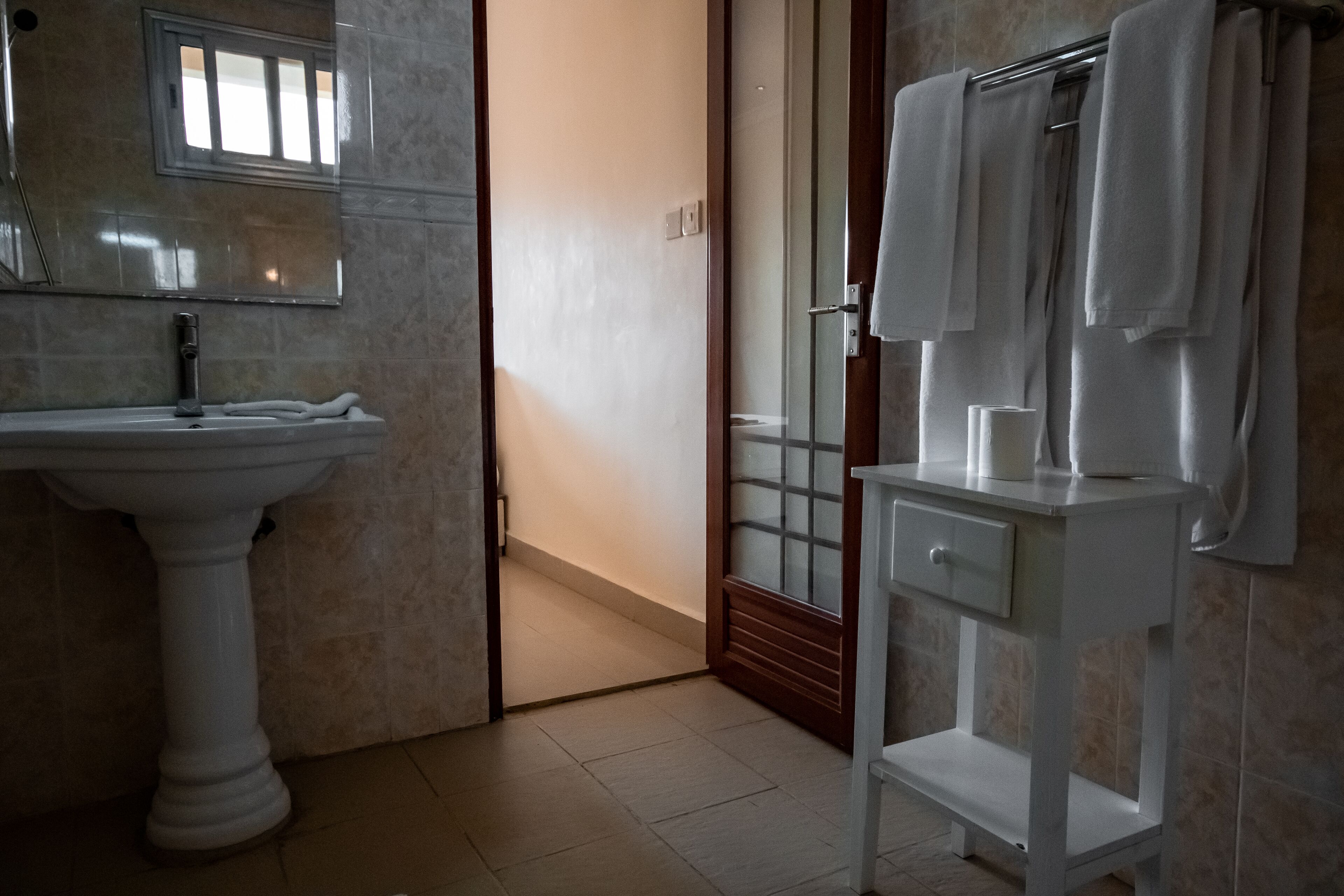 Une salle de bain avec un lavabo sur colonne, un meuble-lavabo avec un miroir et un porte-serviettes, et une porte en verre donnant sur l’espace de la douche.