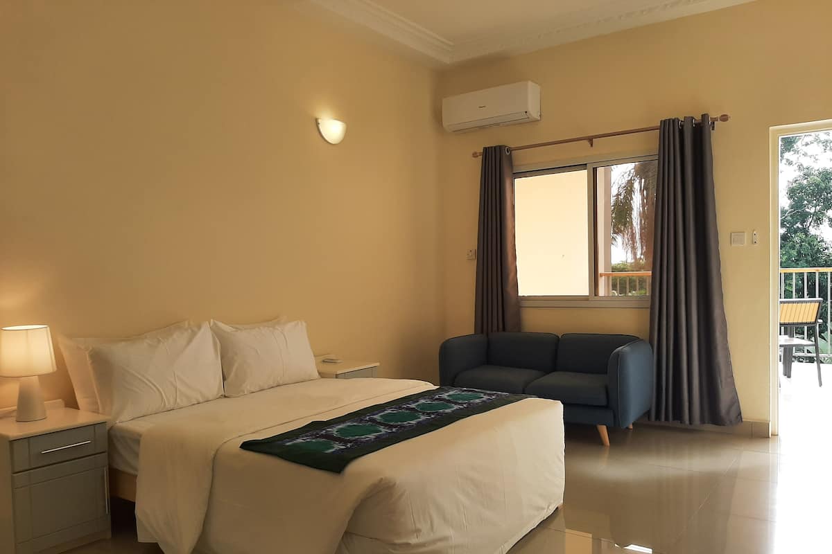 Ein Hotelzimmer mit Bett, Nachttisch, Lampe, Klimaanlage, Fenster mit Vorhängen und einem Balkon mit Stühlen.