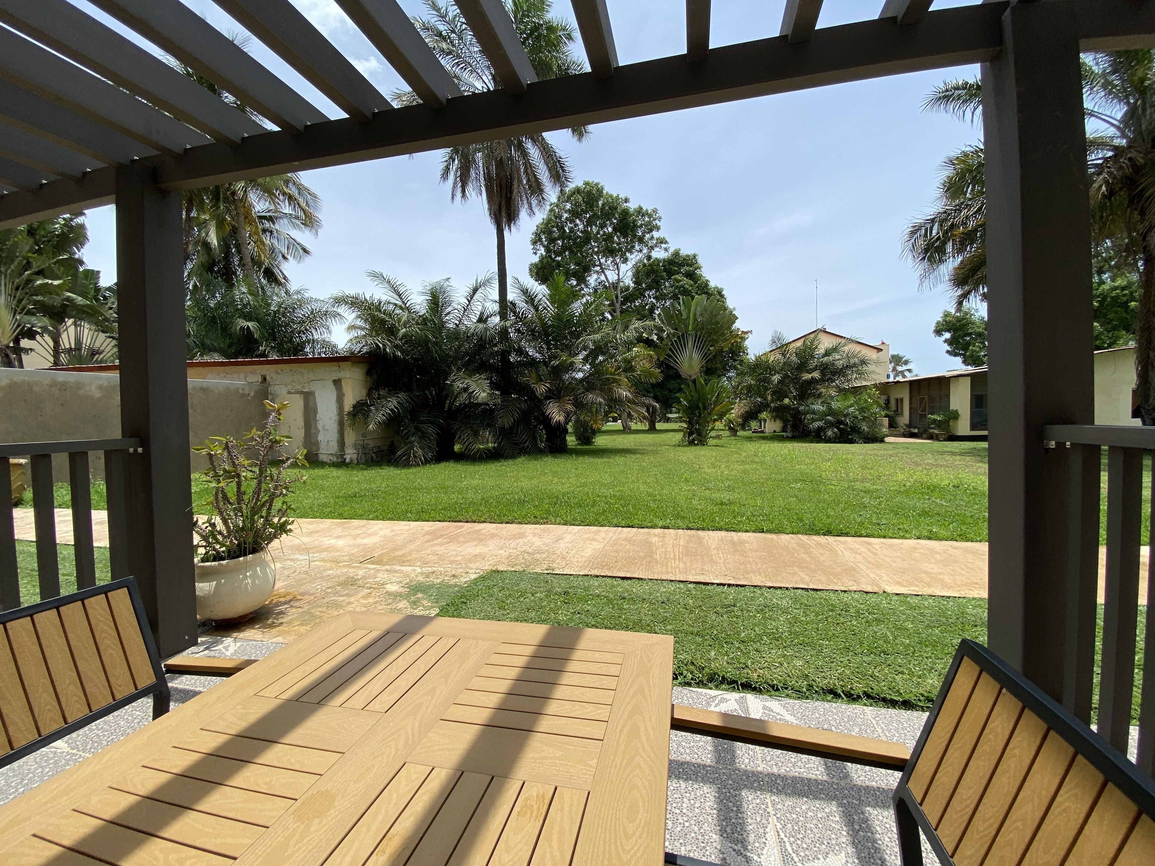 A covered patio with a wooden table and chairs, a concrete pathway, and a well-maintained lawn with palm trees and a building in the background.