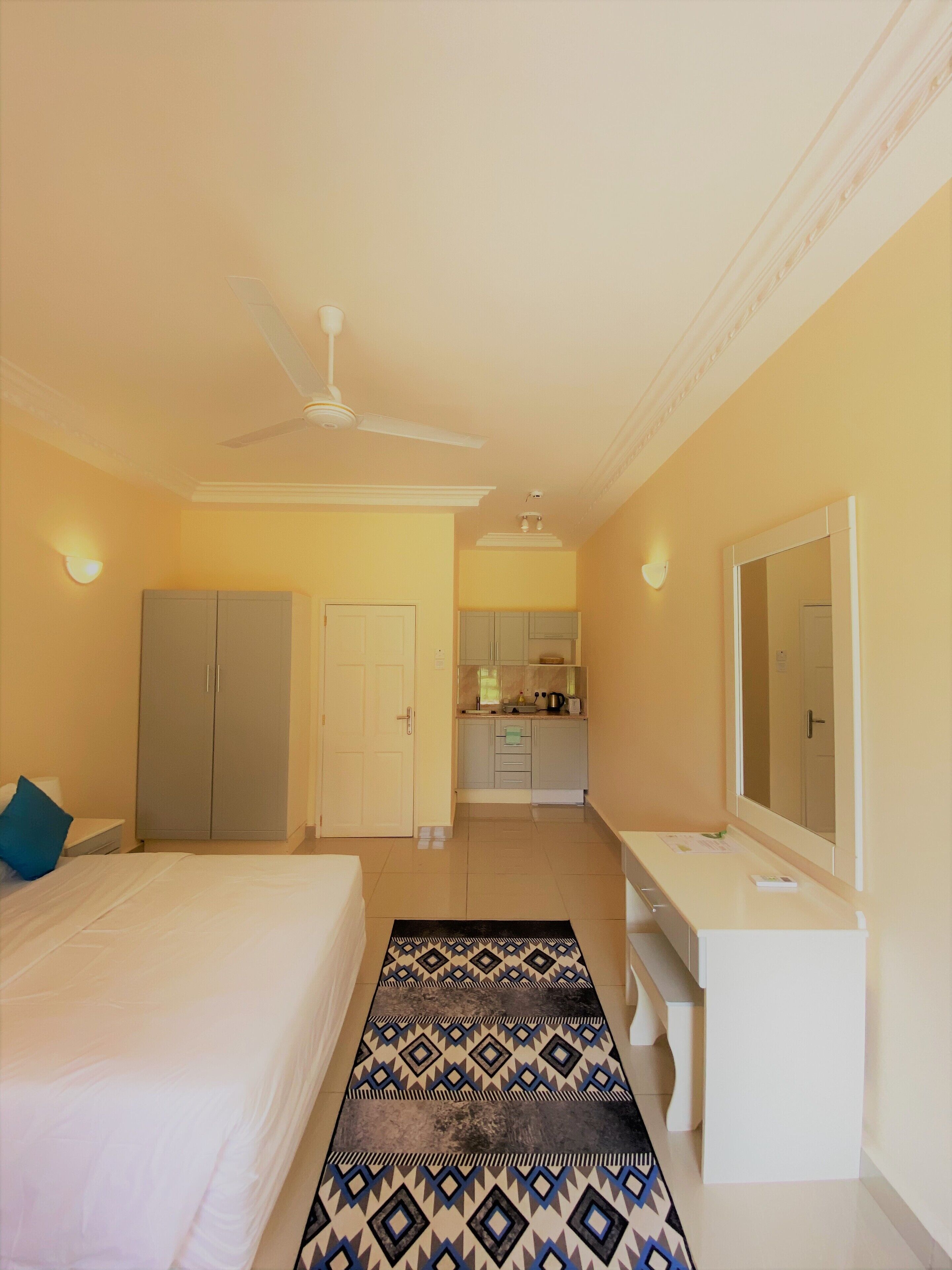 A hotel room with a bed, a desk, a wardrobe, a kitchenette, and a ceiling fan.