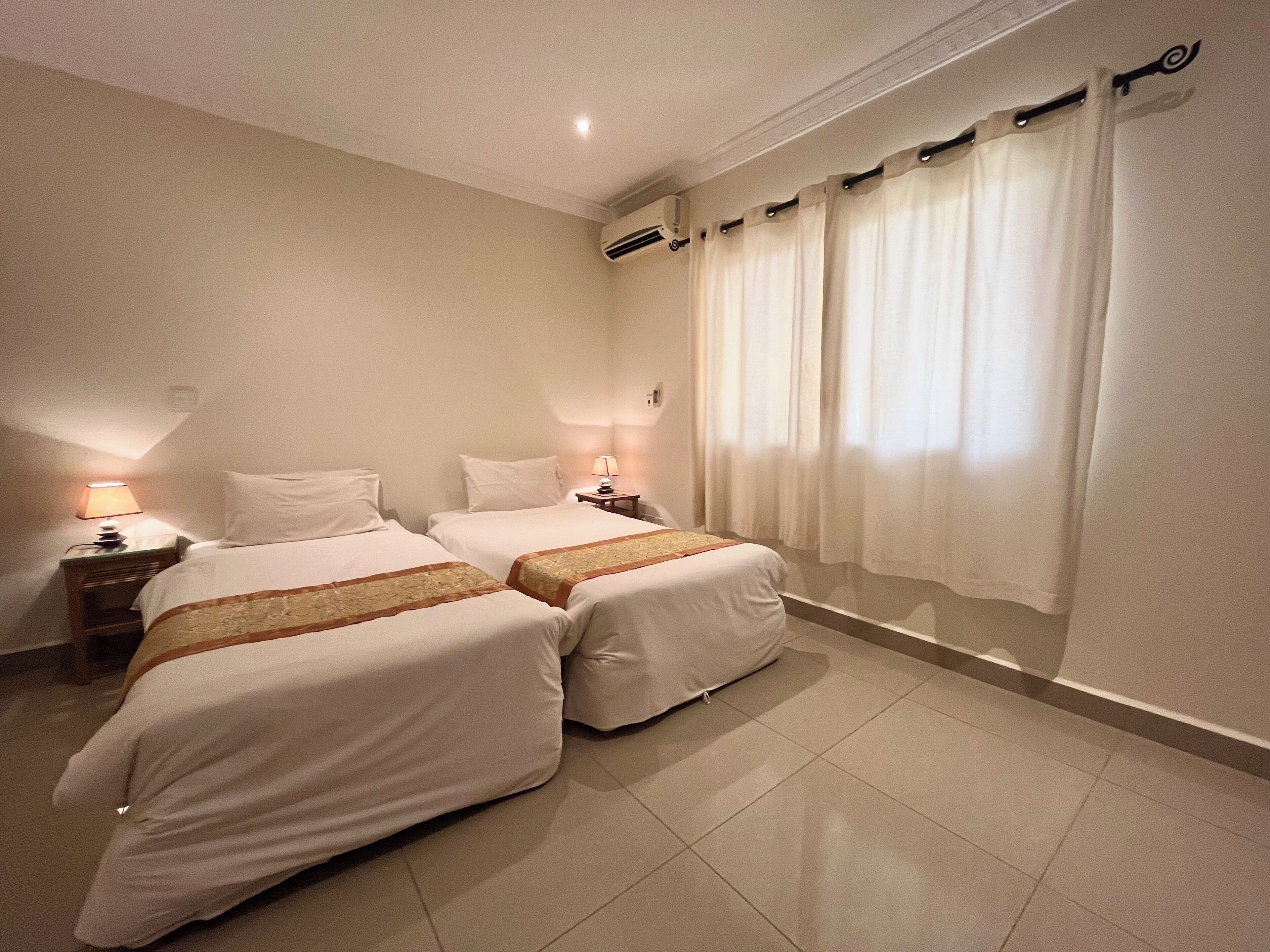 A hotel room with two beds, a nightstand with a lamp, a window with curtains, and an air conditioning unit on the wall.