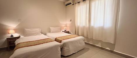 A hotel room with two beds, a nightstand with a lamp, a window with curtains, and an air conditioning unit on the wall.