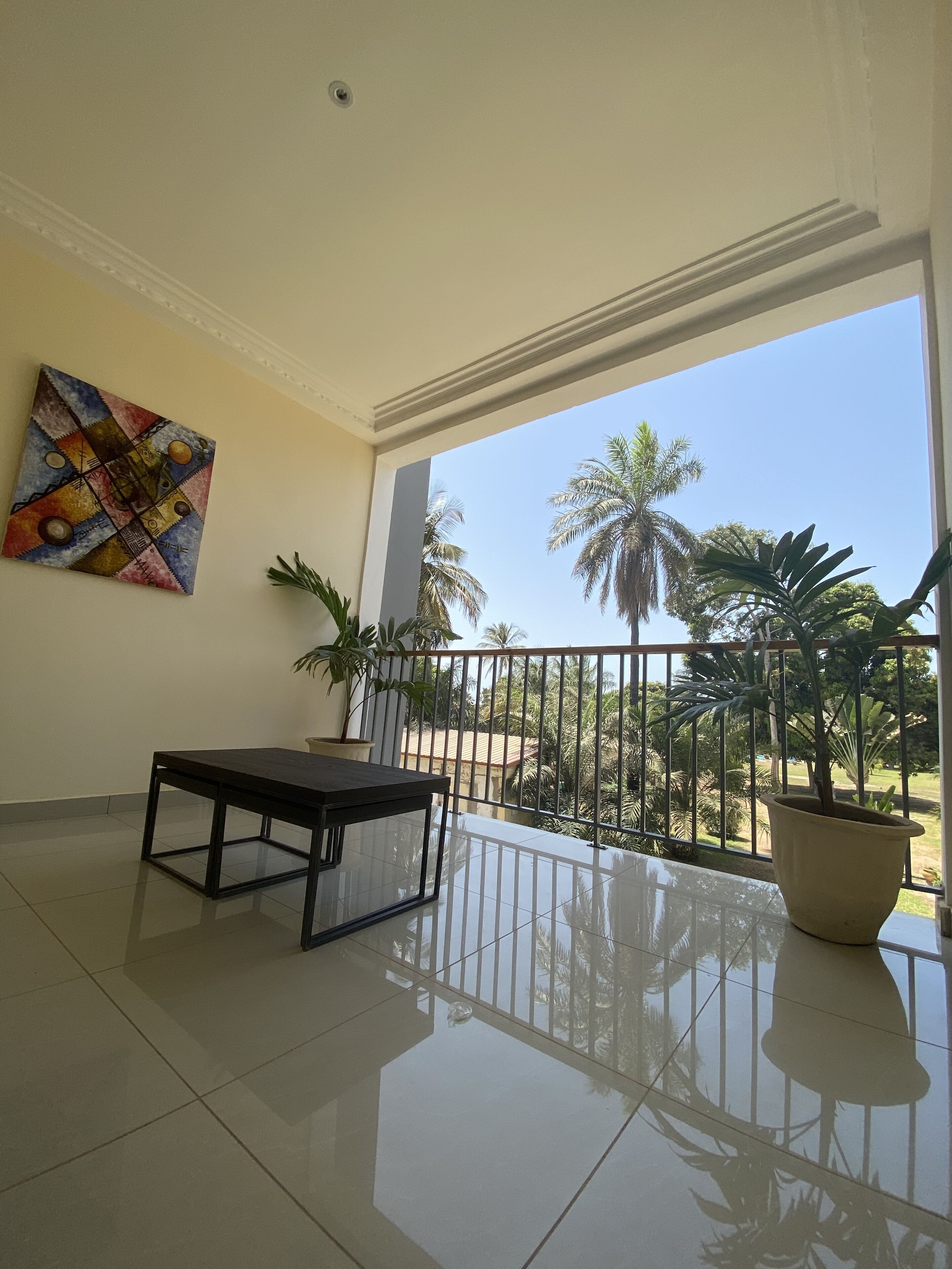 A spacious balcony with a tiled floor, a black metal table, a potted plant, and a colorful abstract painting on the wall.
