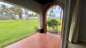 Un porche cubierto con piso de baldosas rojas, un arco de ladrillo y vista a un área verde con palmeras y un edificio al fondo.