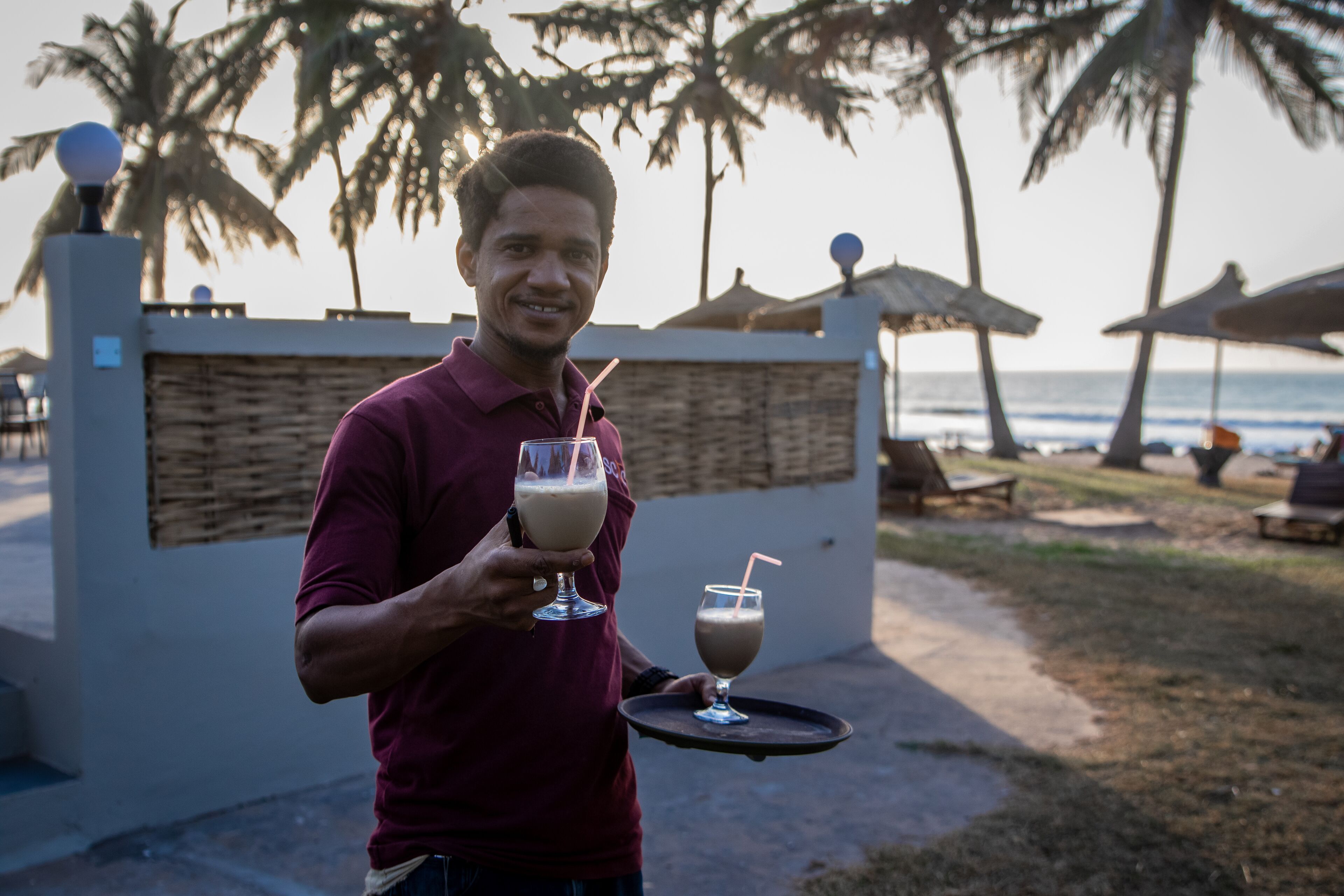 A person holding a tray with two drinks, palm trees, and a beach in the background.