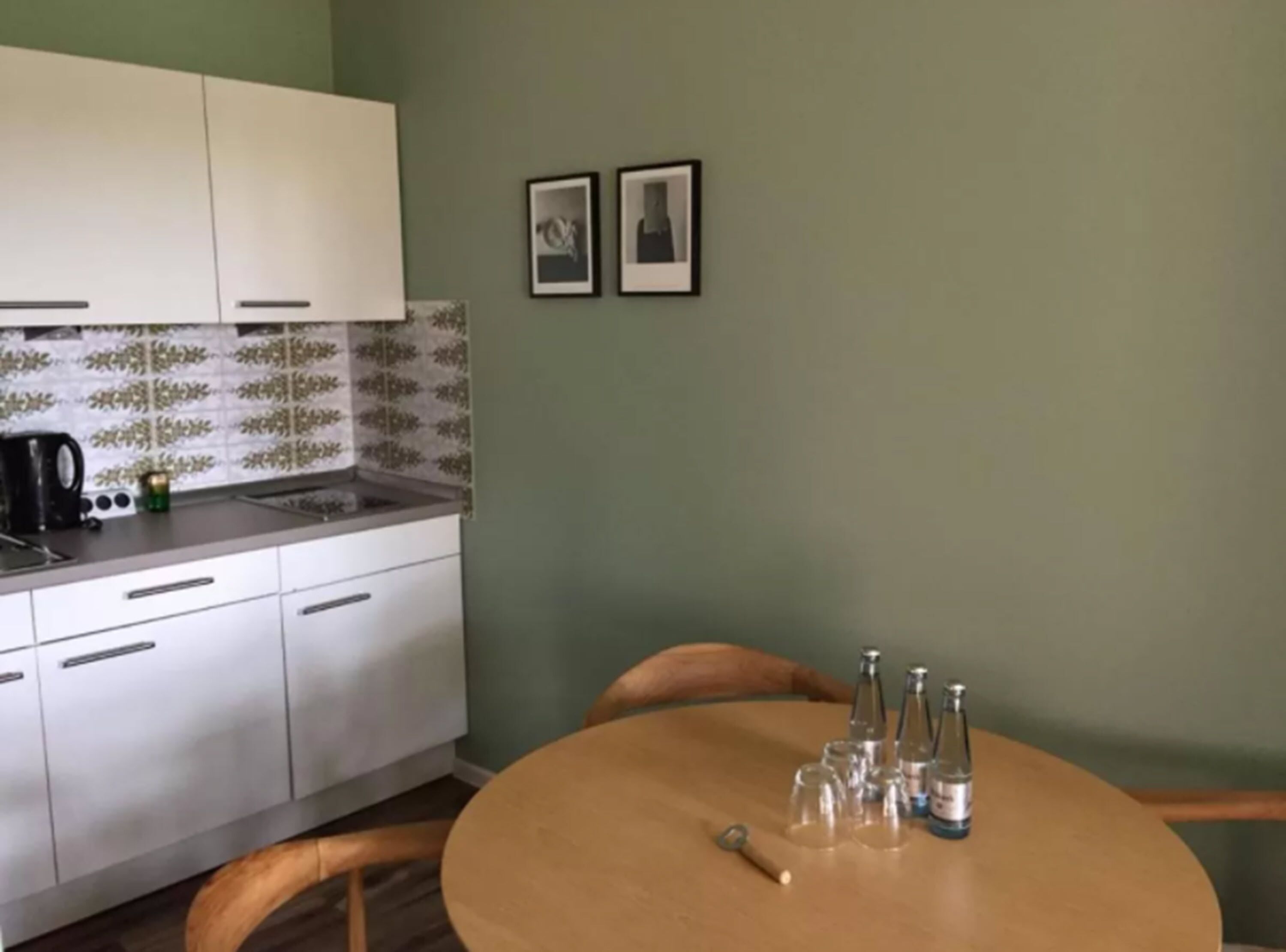 Una cucina compatta con mobili bianchi e paraschizzi piastrellati, un lavandino piccolo e una macchina da caffè. C'è un tavolo da pranzo in legno con sedie e alcune bottiglie e bicchieri sopra.