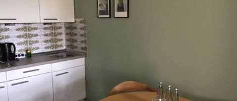 Una cocina compacta con armarios blancos y salpicadero de azulejos, un lavamanos pequeño y una cafetera. Hay una mesa de comedor de madera con sillas y algunas botellas y vasos encima.