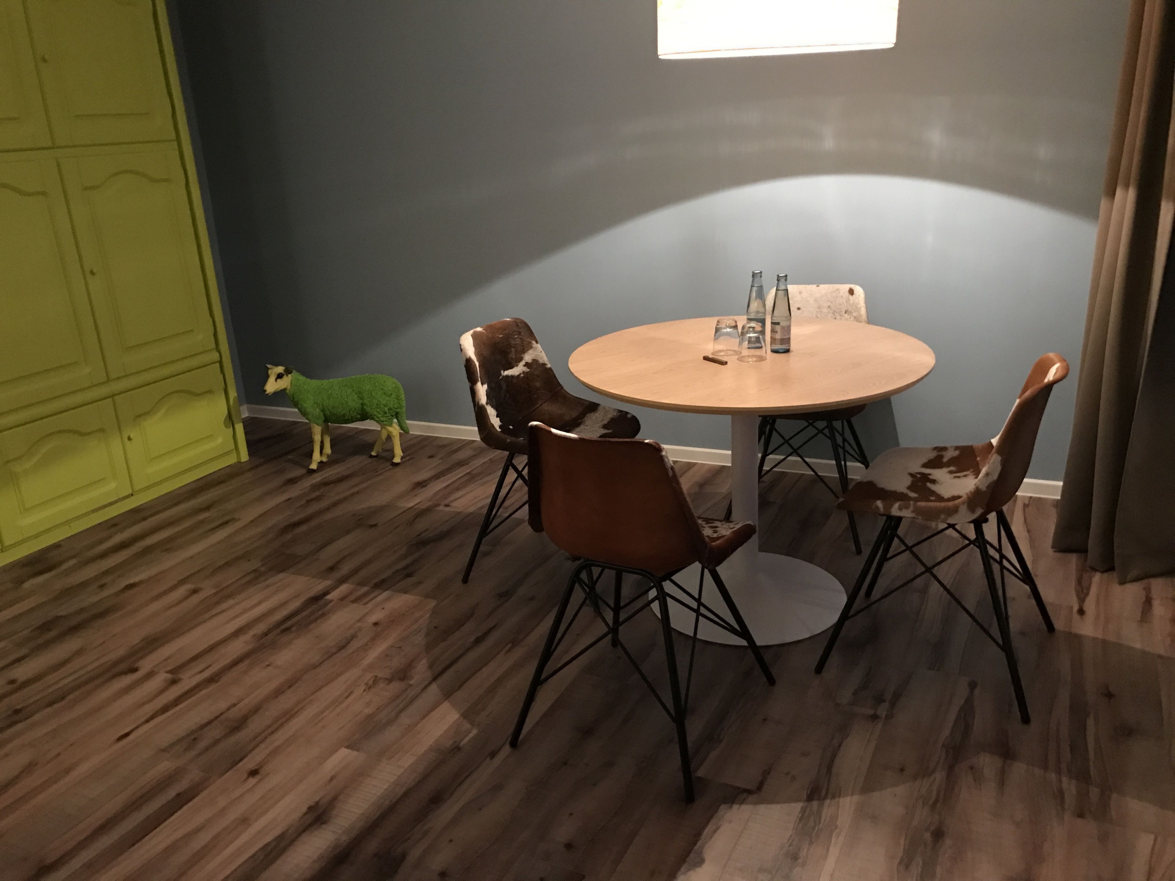 Um quarto com uma mesa redonda de madeira, quatro cadeiras, uma porta verde e uma pequena decoração de ovelha verde.