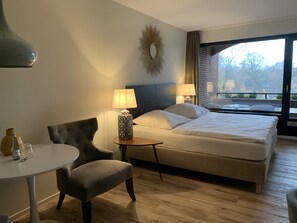 Habitación de hotel con cama, dos lámparas de noche, una mesa redonda con una silla, un adorno en la pared y vistas al exterior.