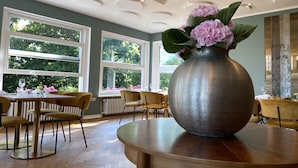 A dining area with a large vase of flowers on a table, multiple chairs, and large windows with a view of greenery. - Das Hotel Ostseeblick (Luetjenburg)