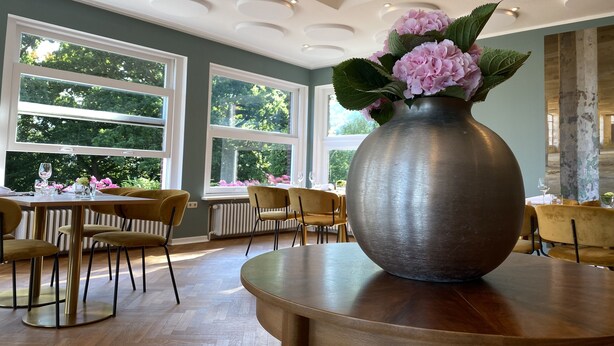 Un espace repas agrémenté d’un grand vase rempli de fleurs posé sur une table, de plusieurs chaises et de grandes fenêtres offrant une vue sur la verdure.