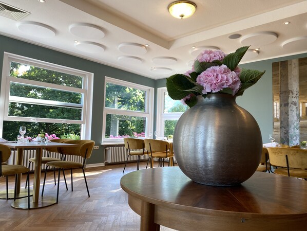 A dining area with a large vase of flowers on a table, multiple chairs, and large windows with a view of greenery. - Das Hotel Ostseeblick (Luetjenburg)