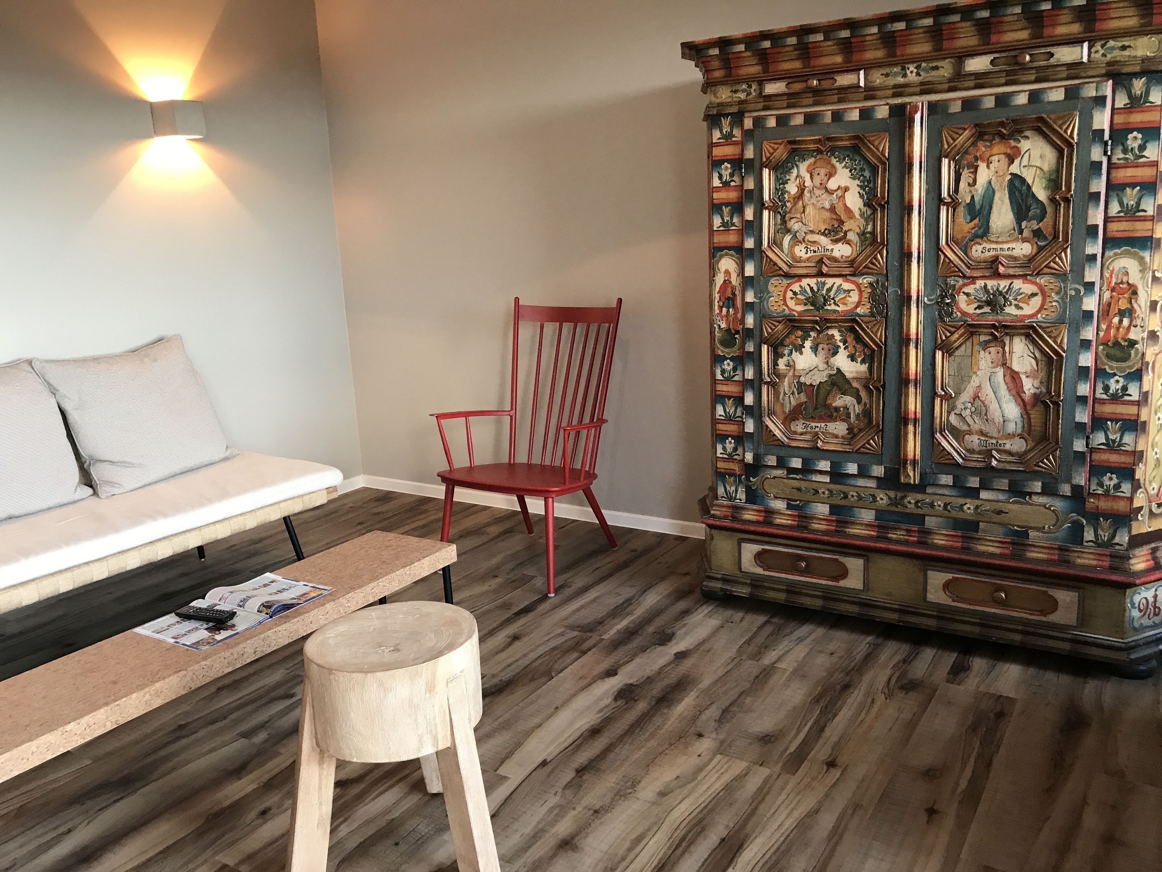 A room with a wooden floor, a red chair, a wooden stool, a bench, and a wall-mounted light fixture.