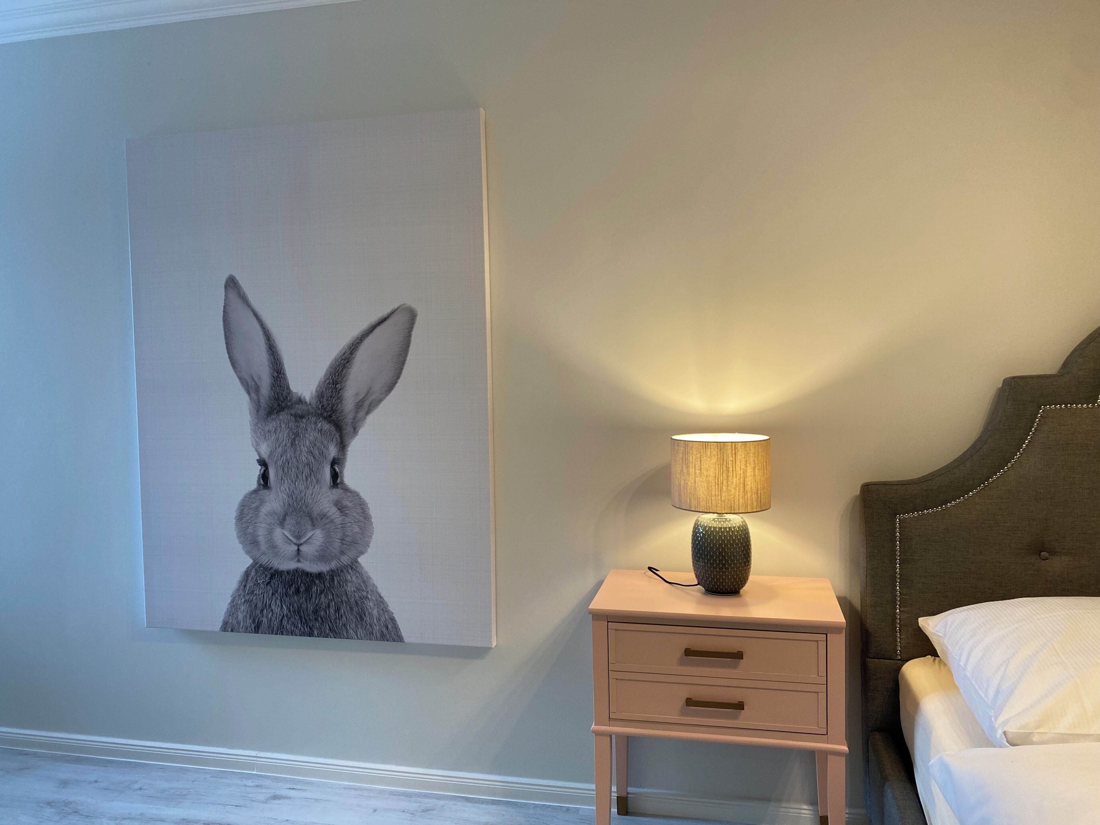 Ein Schlafzimmer mit einem Bett, einem Nachttisch mit Lampe und einem an der Wand hängenden Bild von einem Hasen.