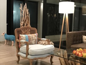 Una sala moderna con una mesa de centro de vidrio, una silla de madera con cojín, un sofá y una lámpara.