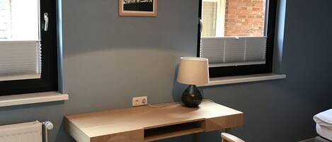 Ett rum med ett skrivbord i trä, en stol, en lampa, ett fönster med persienner och ett element.
