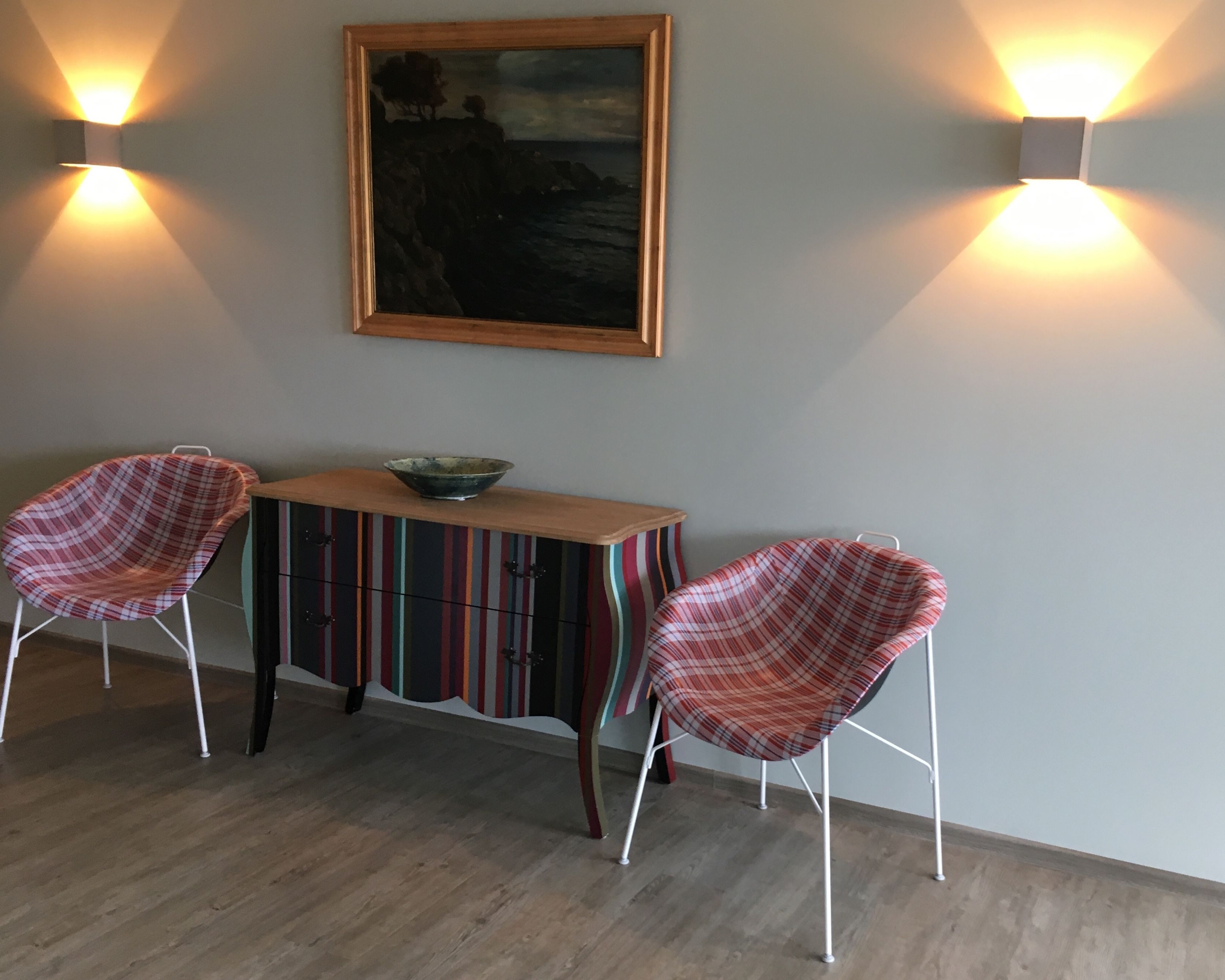 A dining area with two plaid chairs, a wooden table, and a colorful striped cabinet. There are two wall-mounted lights and a framed painting above the table.