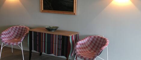 A dining area with two plaid chairs, a wooden table, and a colorful striped cabinet. There are two wall-mounted lights and a framed painting above the table.