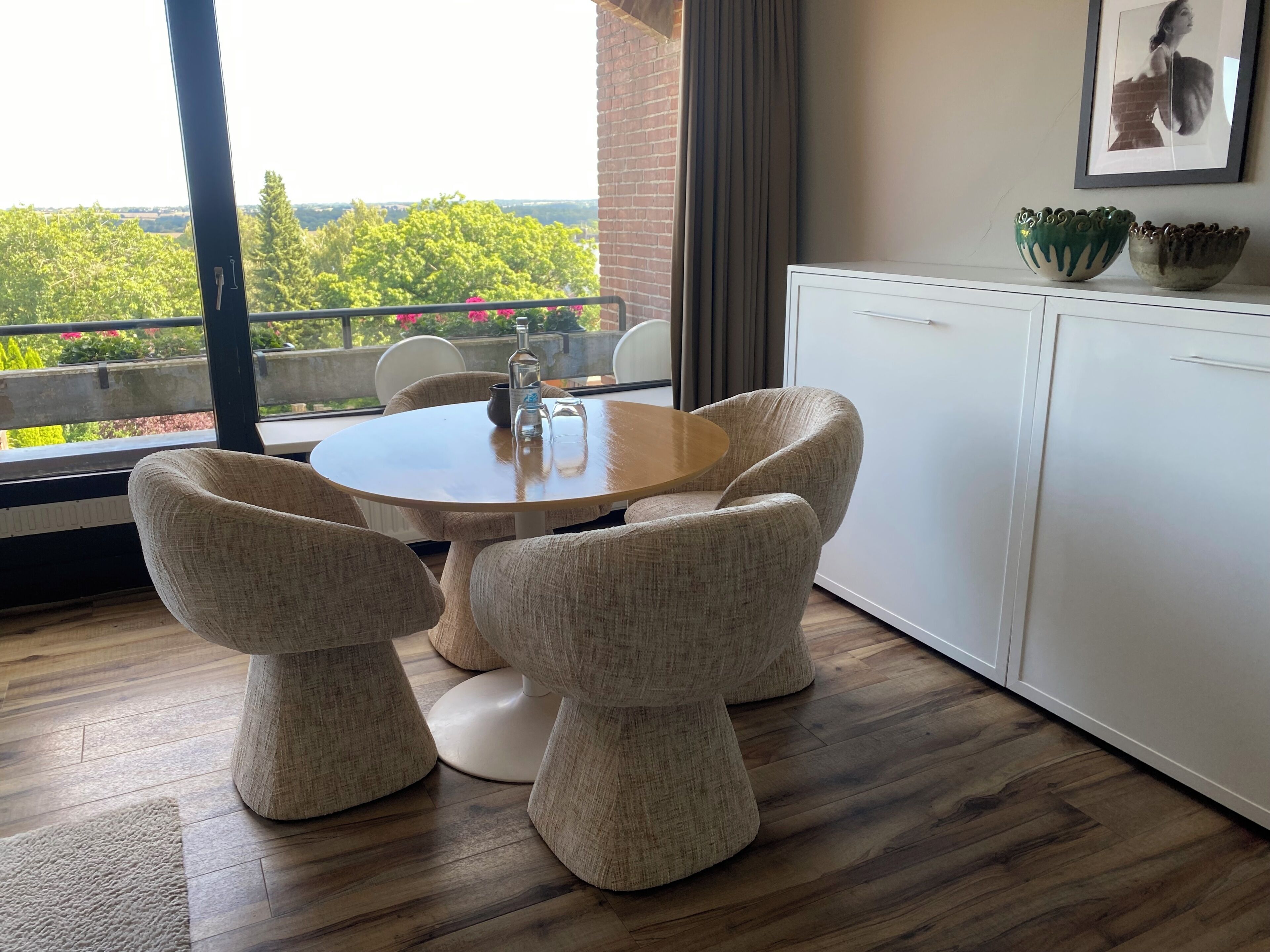 A dining area with a round table and four chairs, a window with a view of trees, and a white cabinet with decorative bowls.