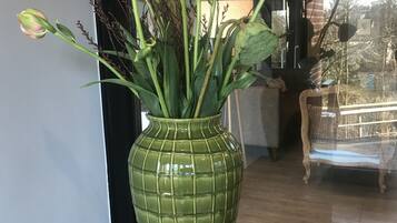 Un vase vert orné de fleurs roses, posé sur un tabouret en bois dans une pièce dotée d’une grande fenêtre et où l’on aperçoit une chaise au fond.