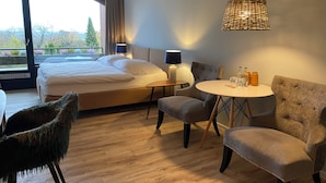 A hotel room with a large bed, a round dining table, two armchairs, a small side table with lamps, and a view of the outdoors. - Das Hotel Ostseeblick (Luetjenburg)