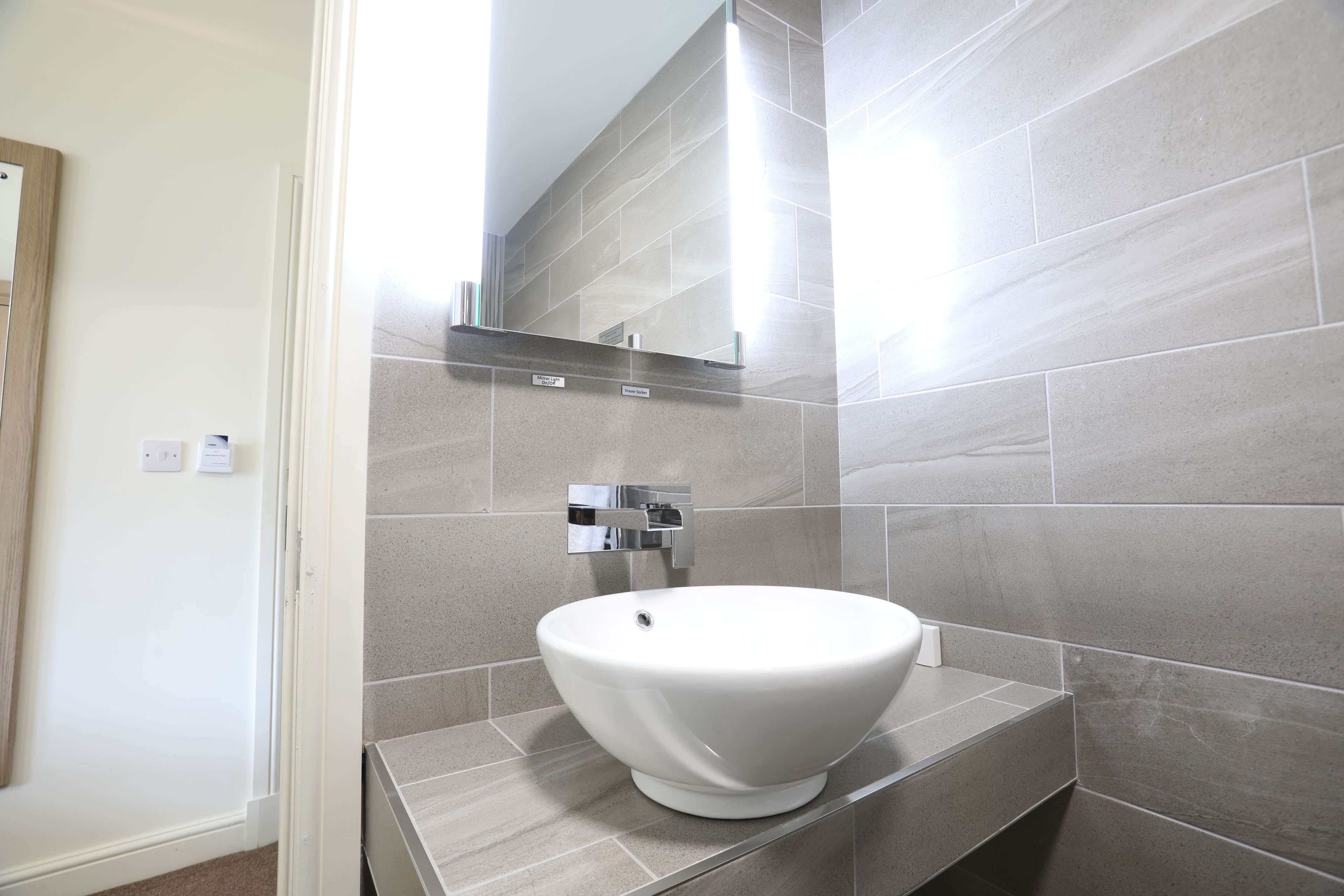 Een moderne badkamer met een wit zuilwastafeltje, een spiegel en grijze betegelde wanden.