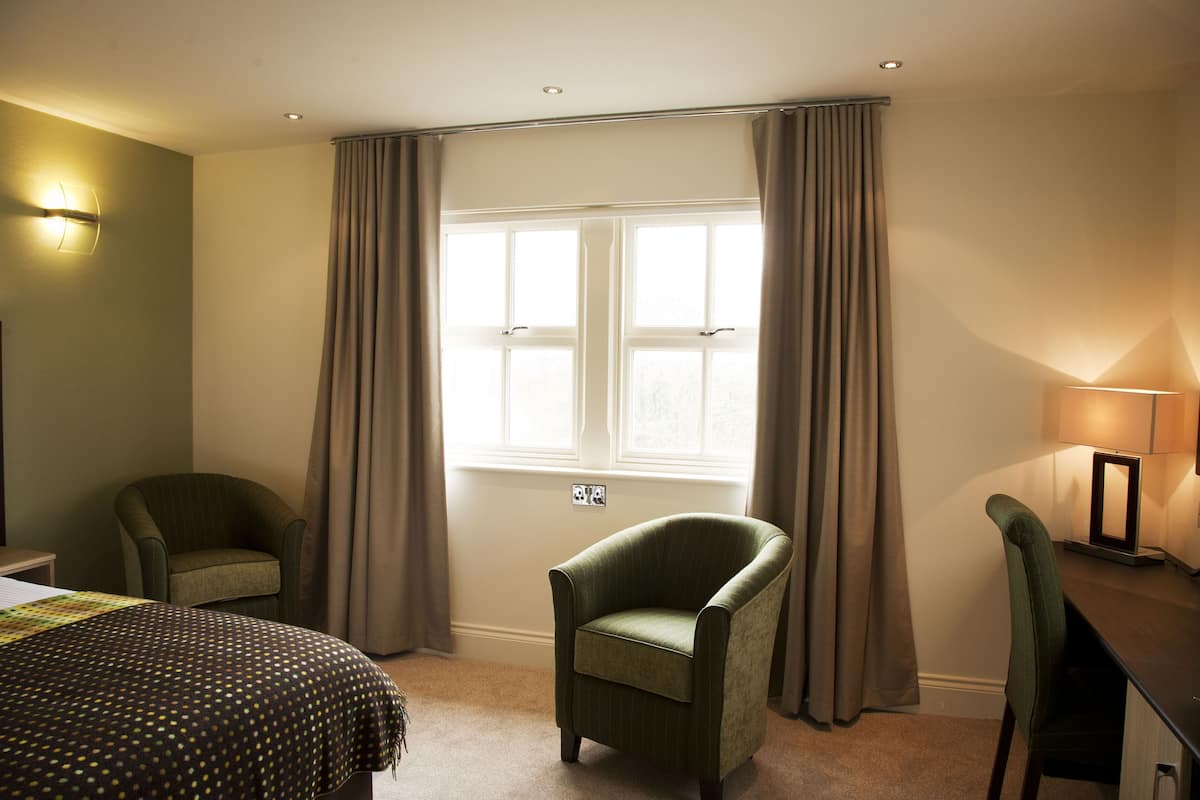 A hotel room with a bed, two chairs, a desk with a lamp, and a window with curtains.