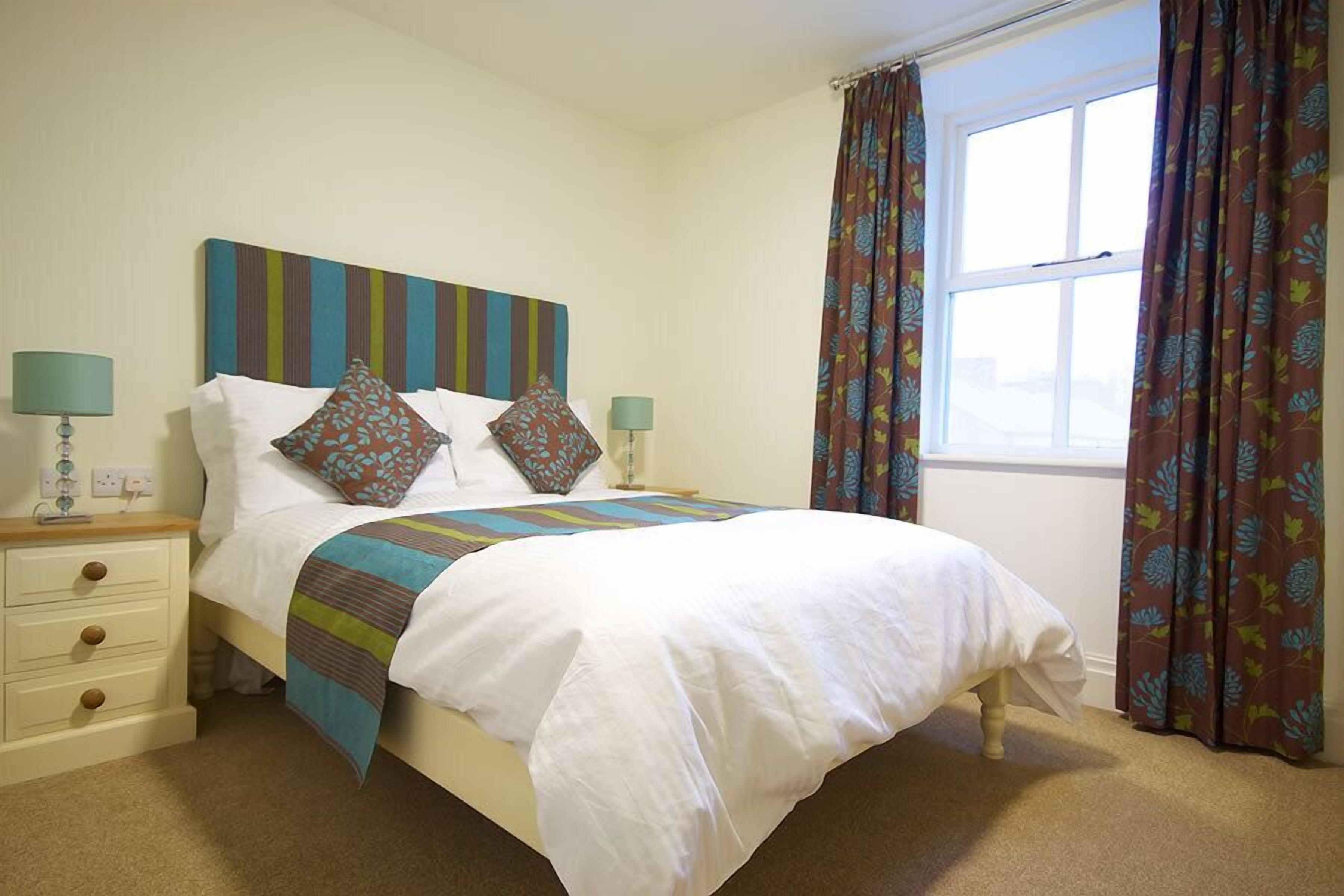 A neatly made bed with a striped headboard, two bedside lamps, and a window with floral curtains.