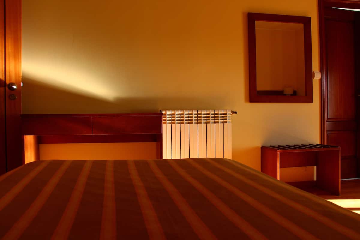 A hotel room with a bed, a radiator, a mirror, and a wooden door.