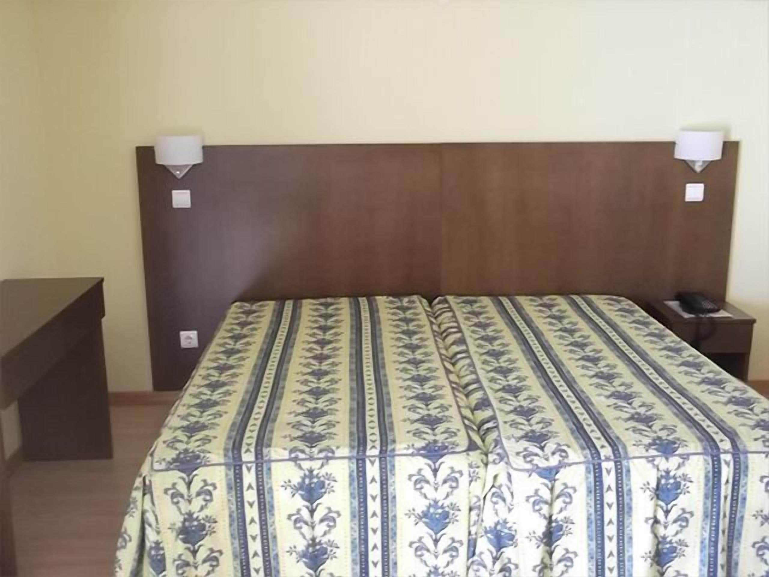Una habitación de hotel con cabecera de madera, colcha estampada, mesita de noche con lámpara y teléfono.