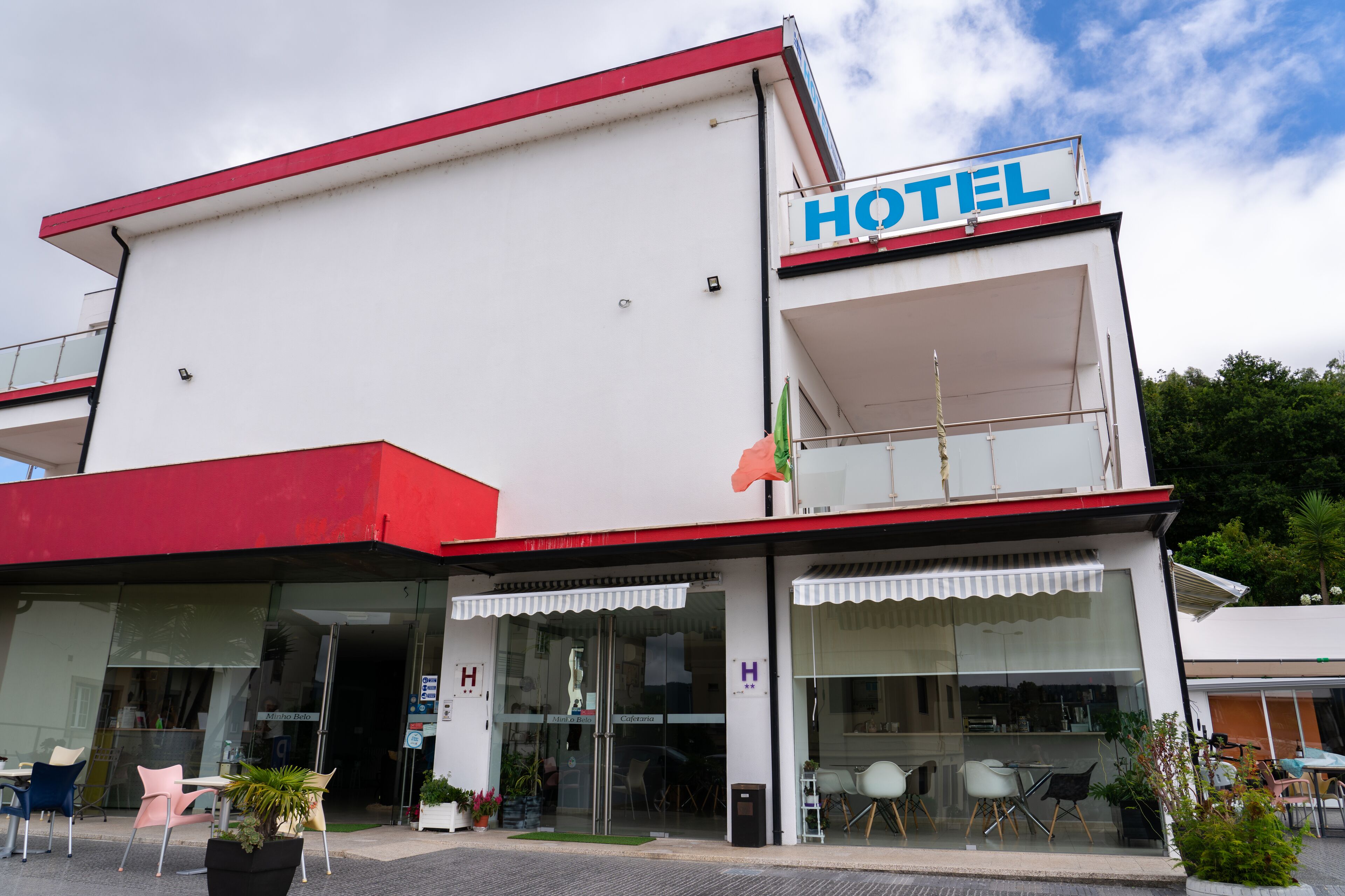 Un hôtel à la façade rouge et blanche, doté de grandes fenêtres en verre et d’un espace extérieur aménagé pour s’asseoir.