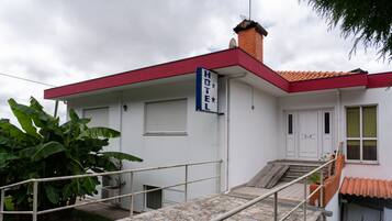 A white building with a red roof and a sign that reads "HOTEL". There is a staircase leading to the entrance, and a large plant is visible to the left.