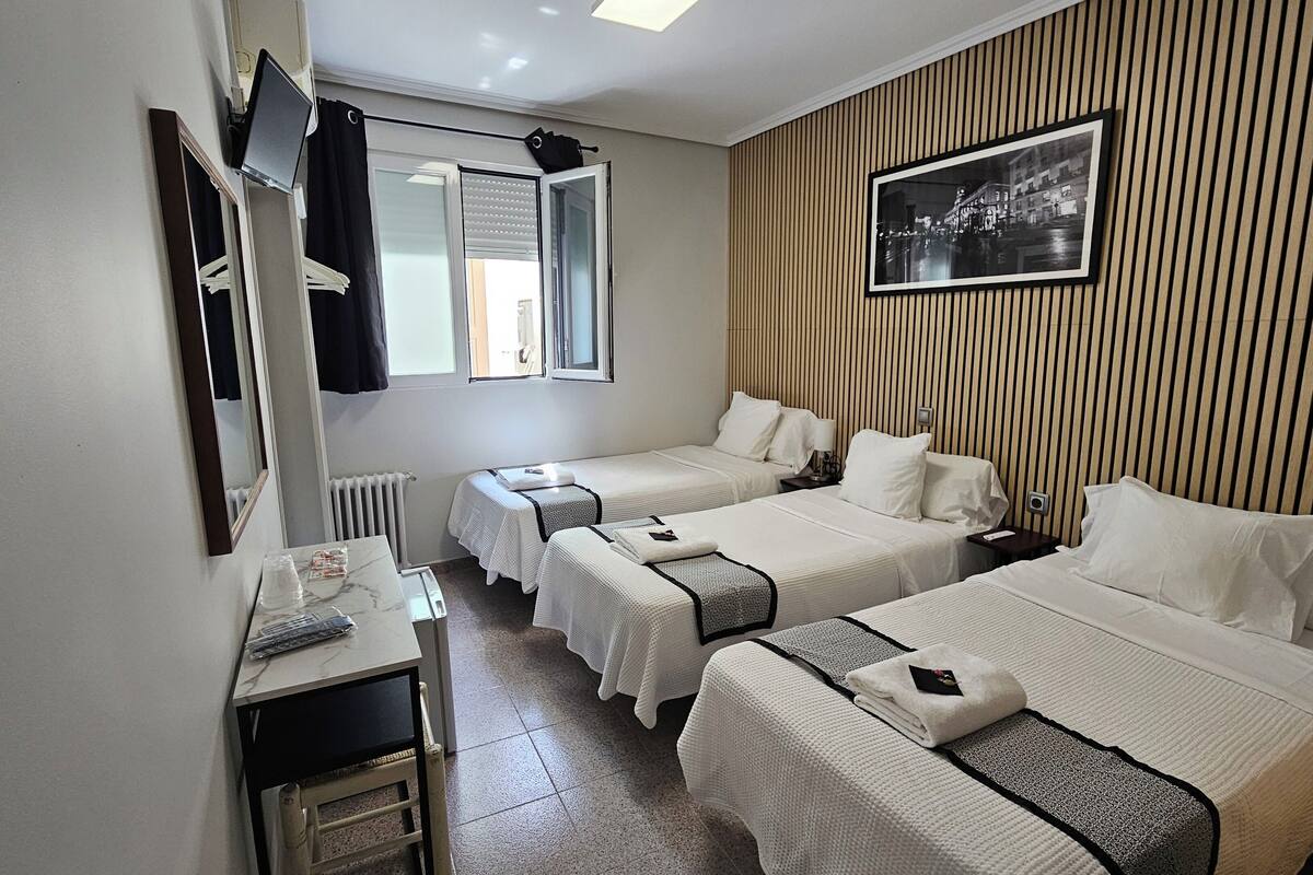 A hotel room with two beds, a desk, a mirror, a window with blinds, a radiator, and a framed picture on the wall.
