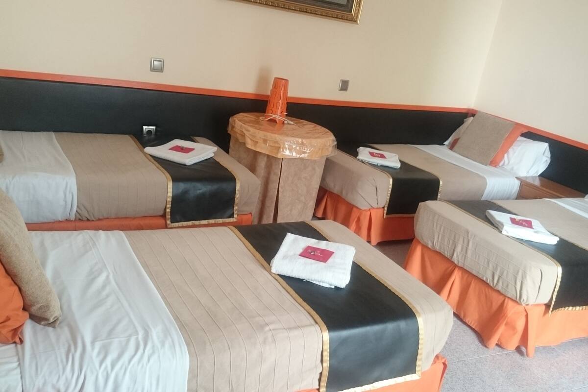 A hotel room with four single beds arranged in a corner, each with a pillow and a folded towel. There is a small round table with a lamp between the beds.