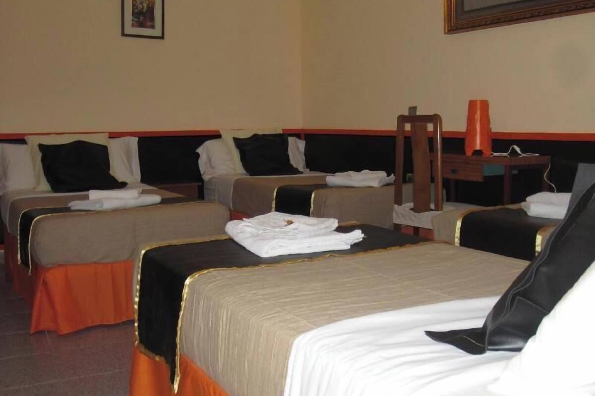 A hotel room with four beds, a wooden chair, a table with a lamp, and a framed picture on the wall.