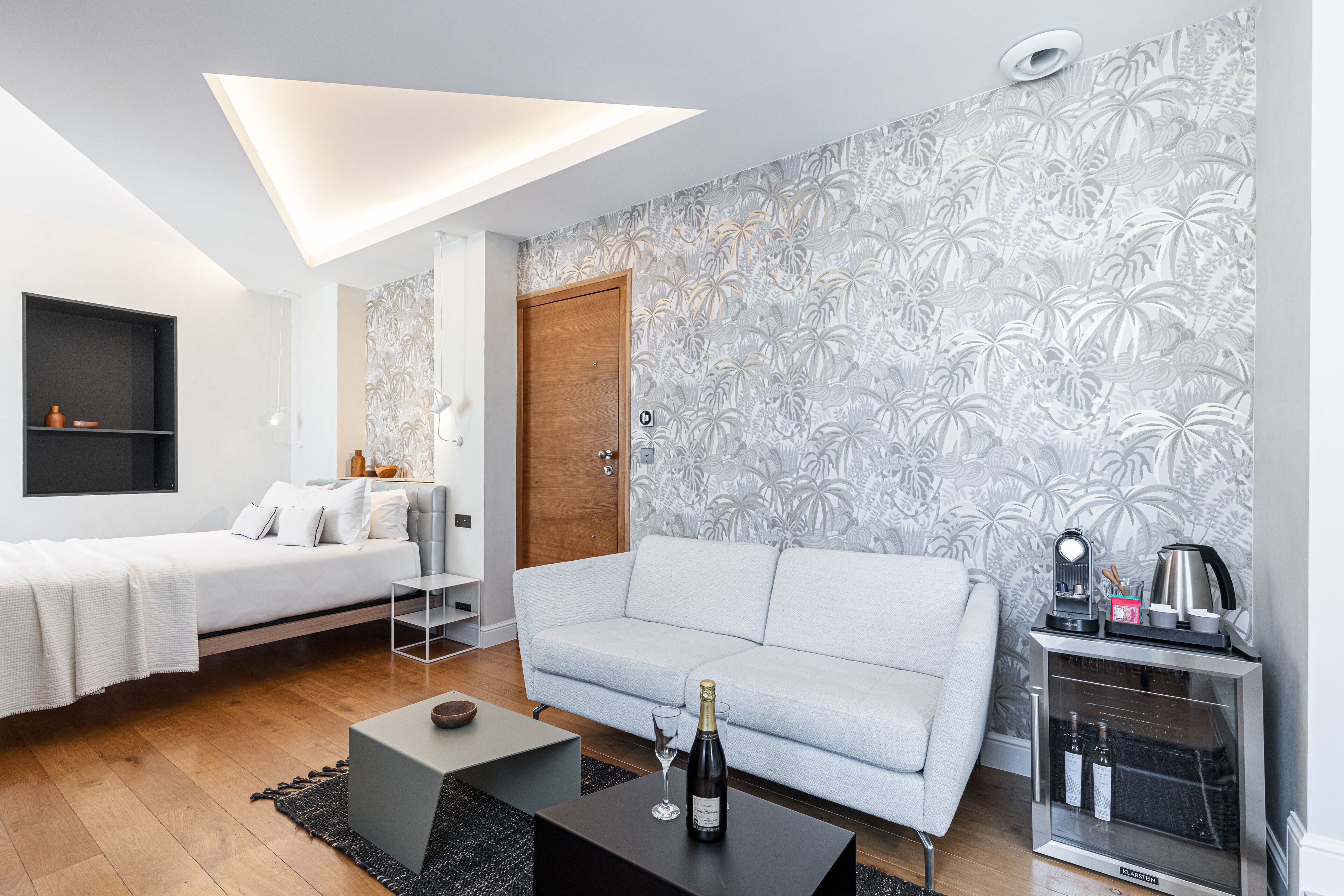 A modern hotel room with a bed, a sofa, a coffee table, and a minibar.
