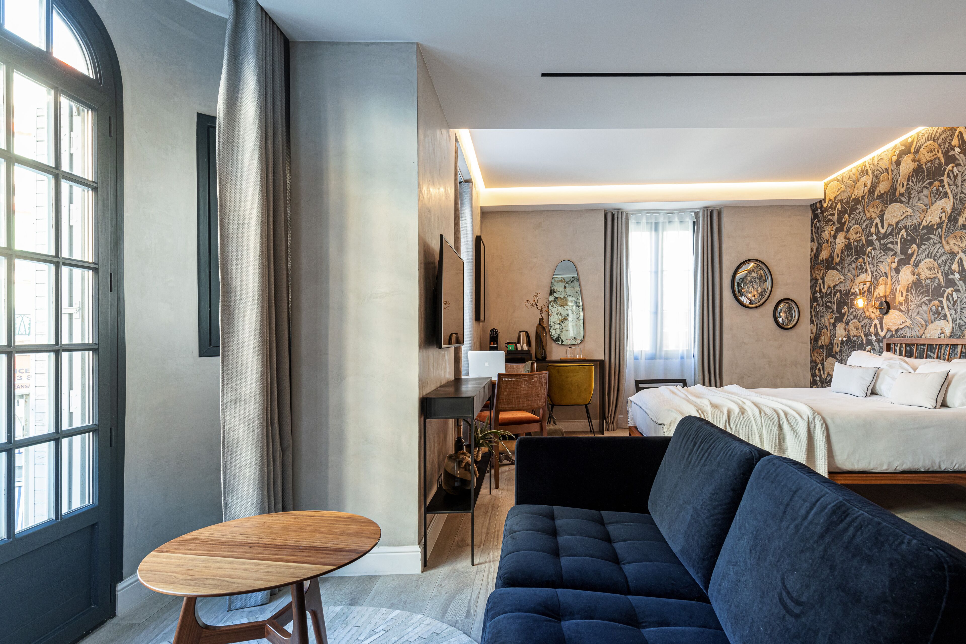 A modern hotel room with a large bed, a dark blue sofa, a wooden table, and a desk with a chair. There is a large window on one side and a patterned wallpaper on the other.