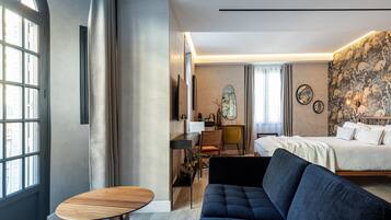 A modern hotel room with a large bed, a dark blue sofa, a wooden table, and a desk with a chair. There is a large window on one side and a patterned wallpaper on the other.
