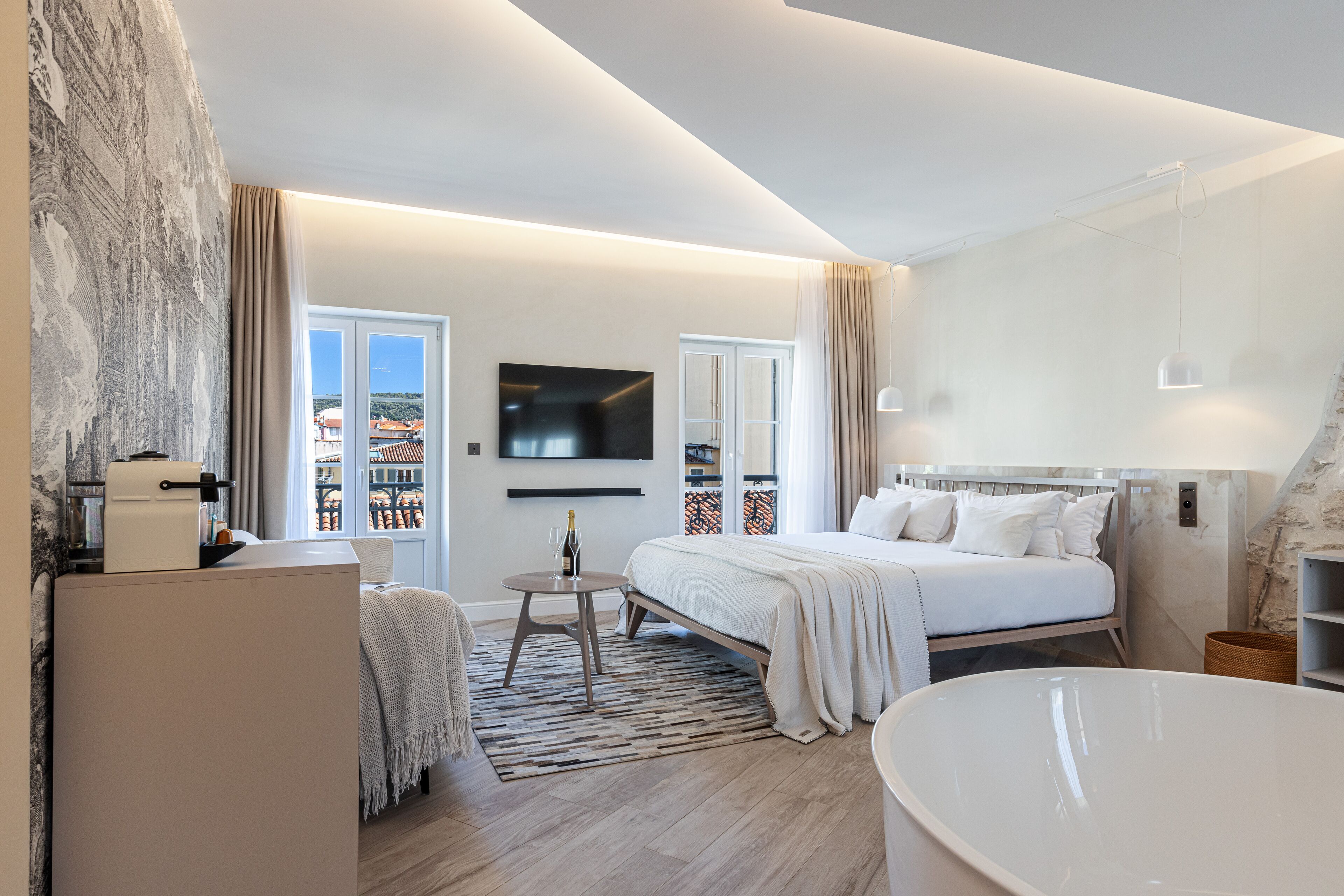 Una camera d'albergo moderna con un letto grande, una zona salotto con un tavolino, una TV a schermo piatto montata a parete e vista sulla città dalle finestre.