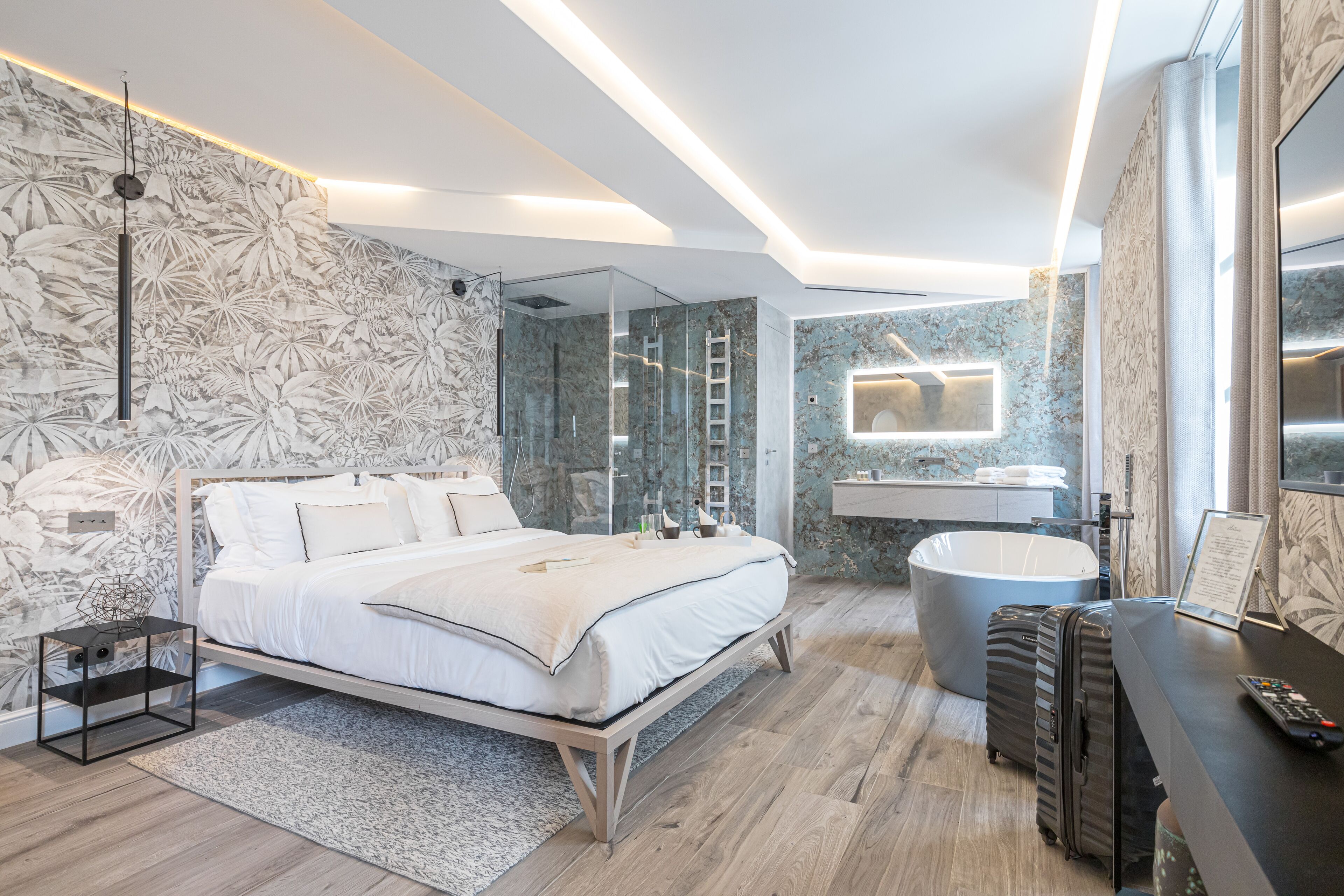 A modern bedroom with a large bed, a glass-enclosed shower, a vanity with a mirror, and a desk with a suitcase.