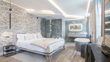 A modern bedroom with a large bed, a glass-enclosed shower, a vanity with a mirror, and a desk with a suitcase.