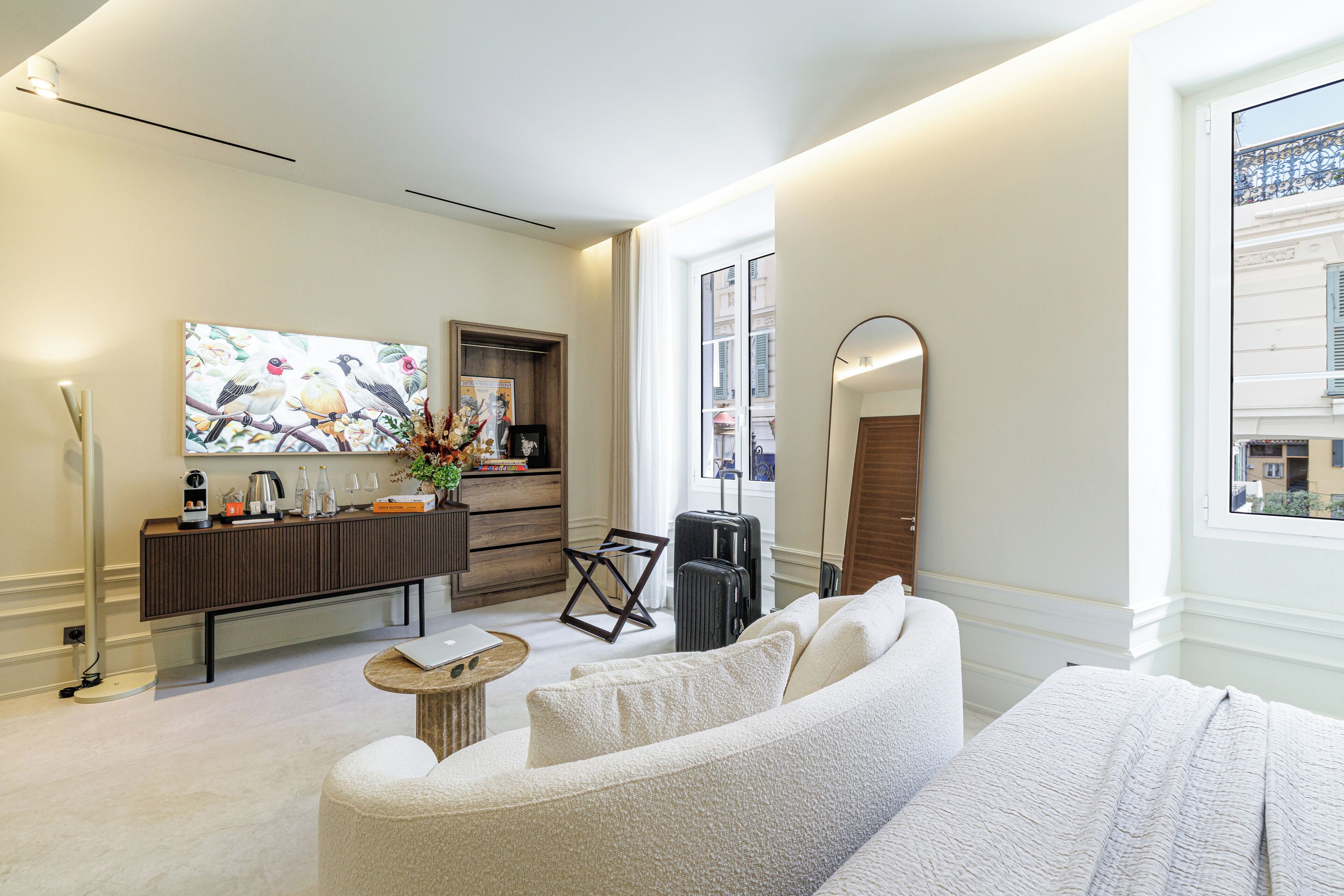 Una habitación de hotel moderna con sofá, una mesita redonda, cómoda, espejo y ventana con vistas a edificios.
