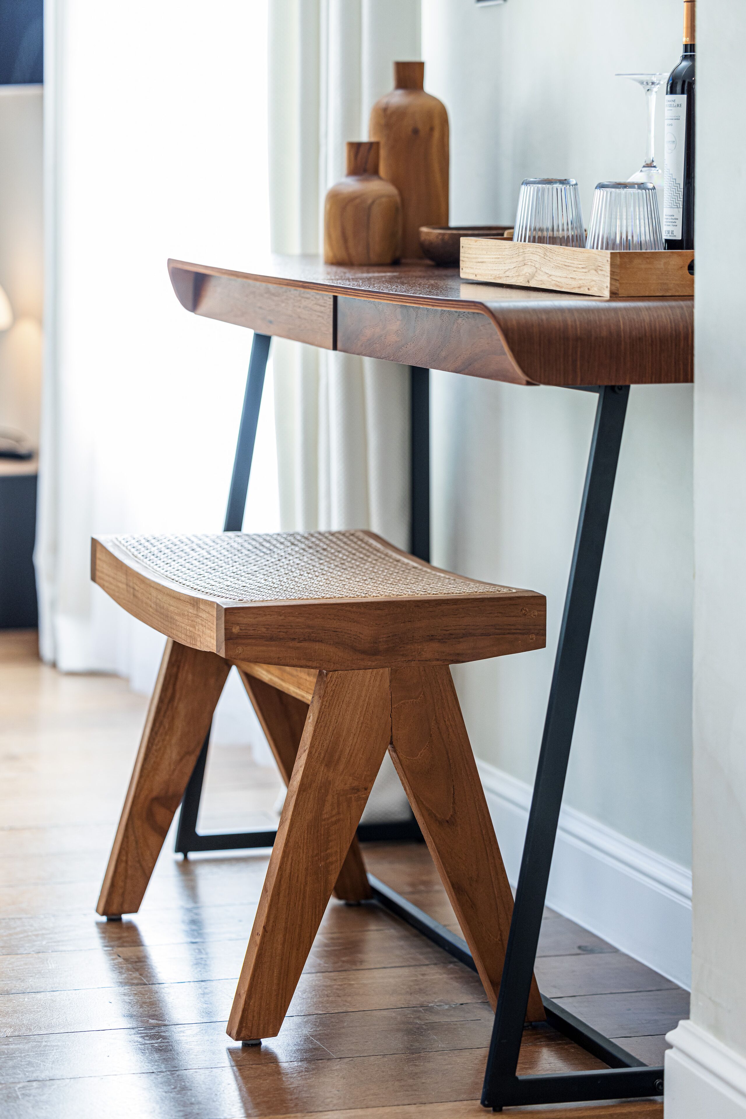 A wooden bench with a woven seat and metal legs, placed on a wooden floor next to a table with a glass tray and wine glasses.