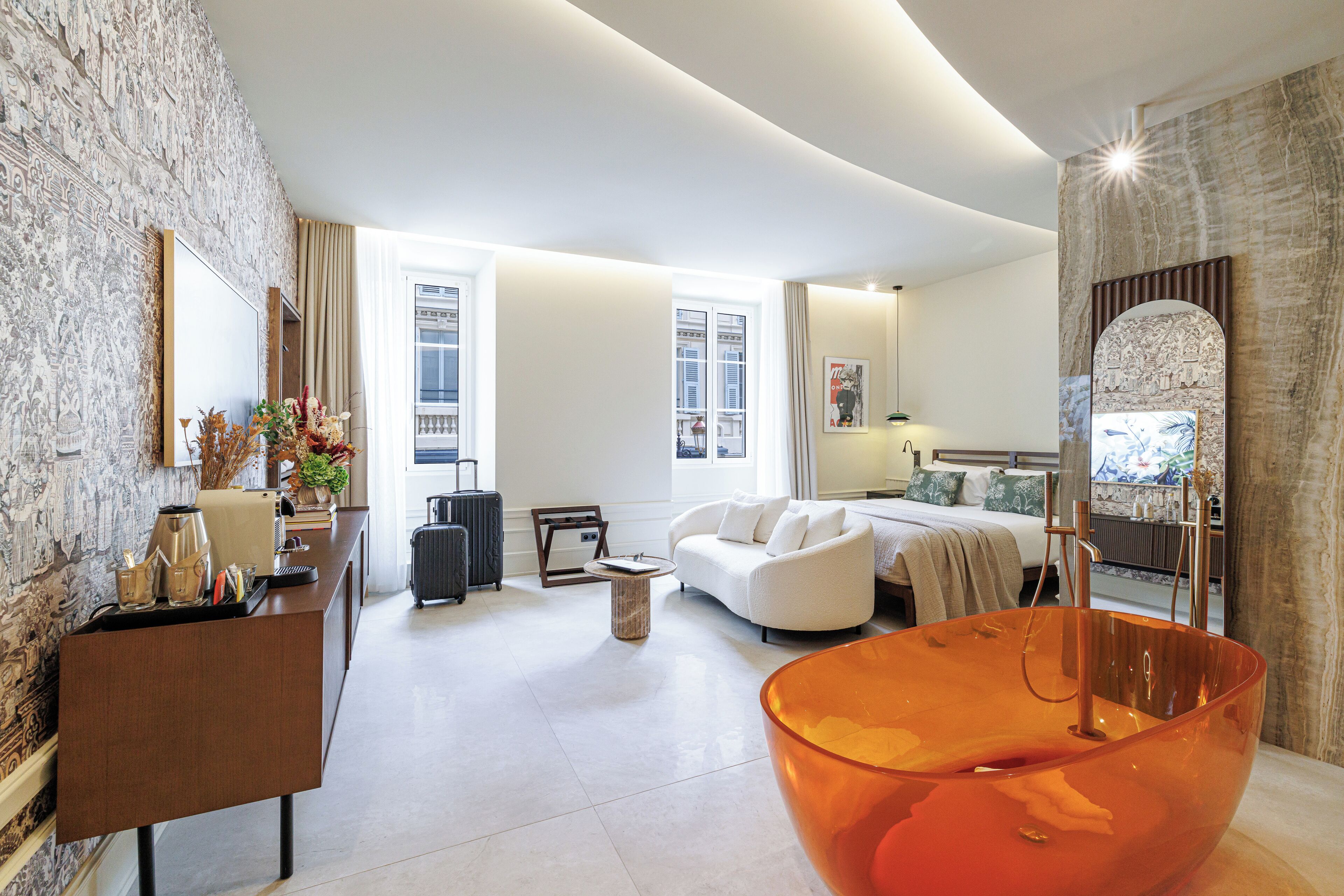 A modern hotel room with a large bed, a sitting area with a sofa, a dining table, and a large orange vase.
