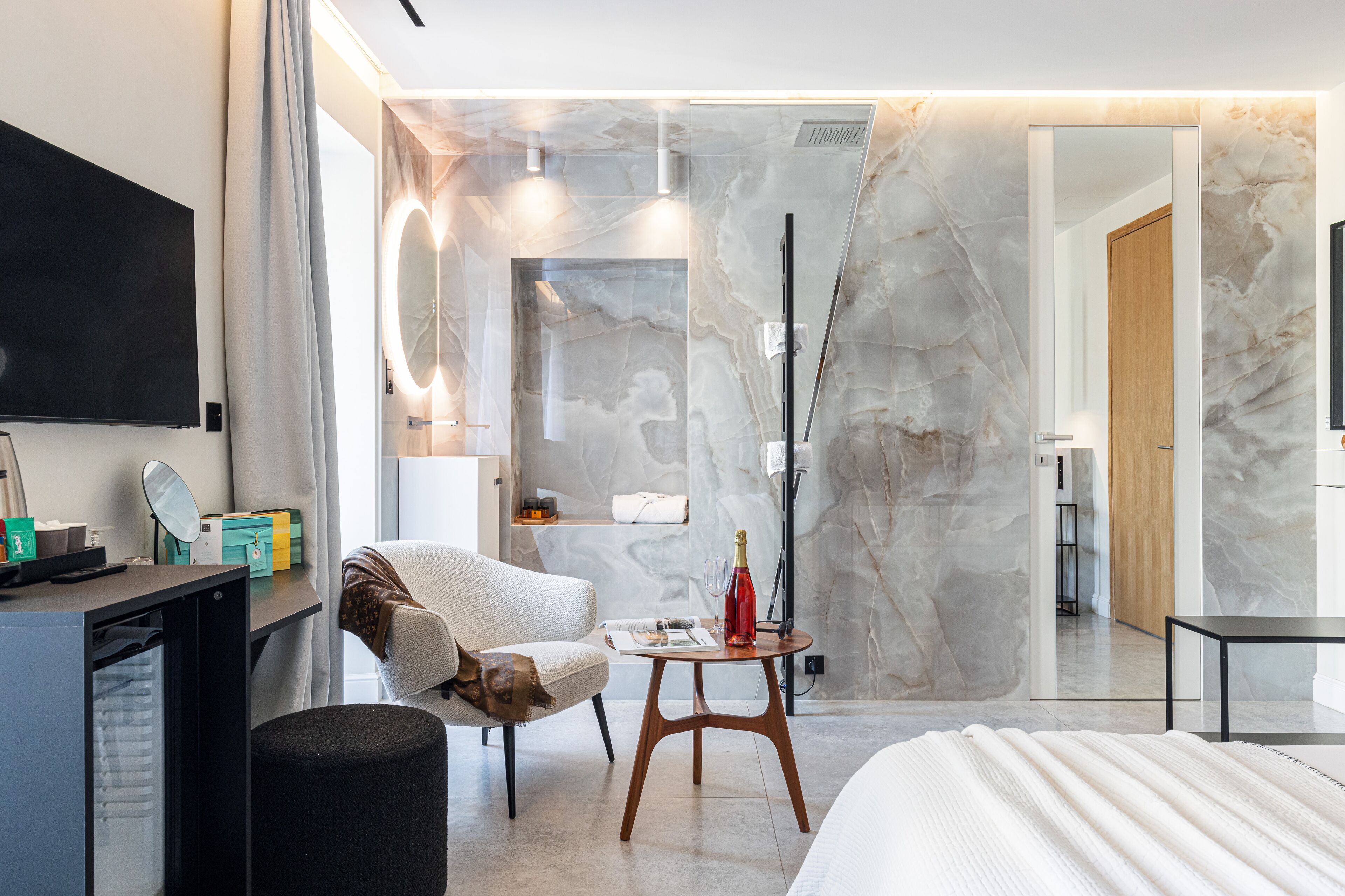 A modern hotel room with a bed, a desk with a computer, a chair, a small table with a bottle, and a mirror.