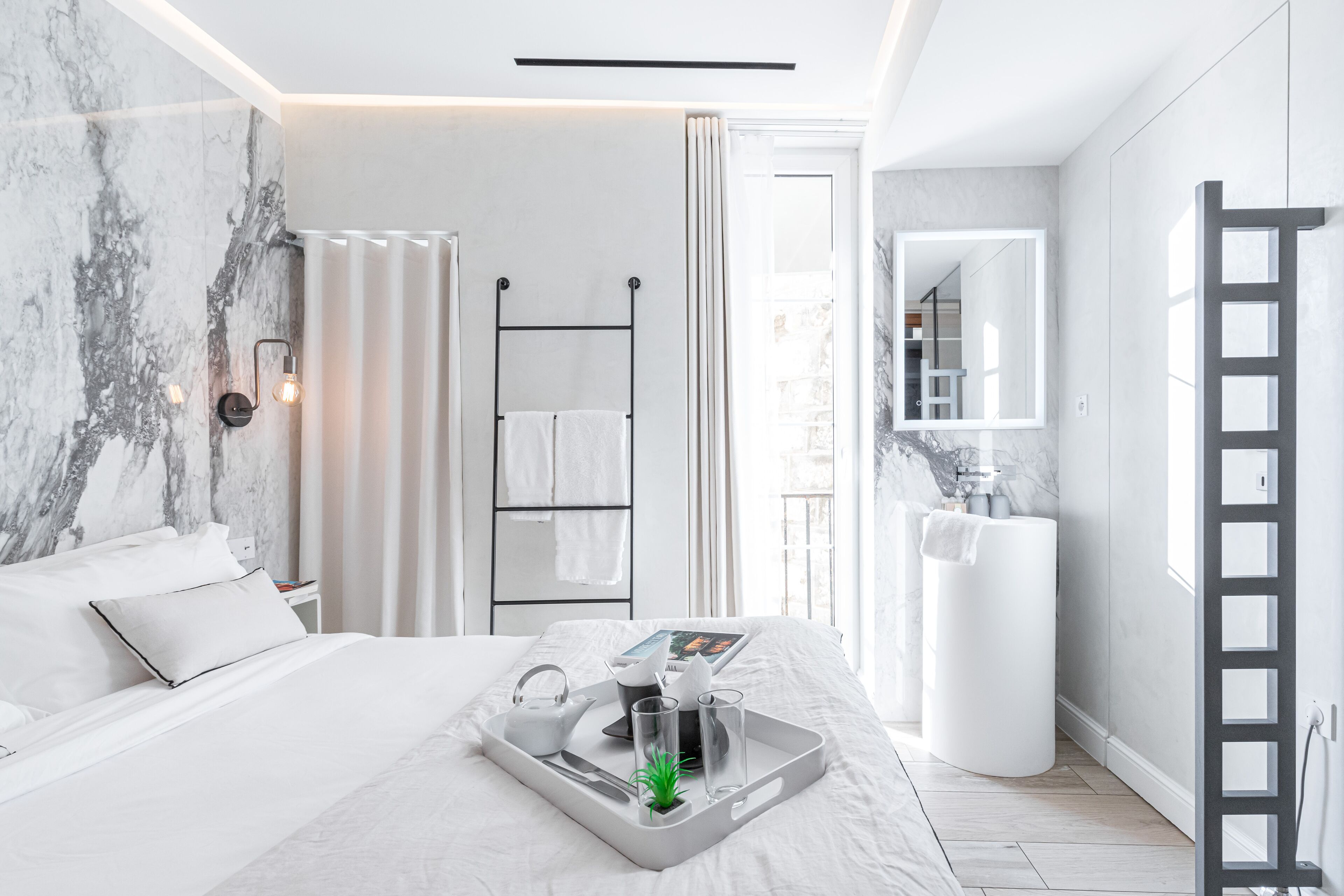 A modern hotel room with a marble wall, a bed with white linens, a bedside table with a tray, a towel rack, and a large mirror.