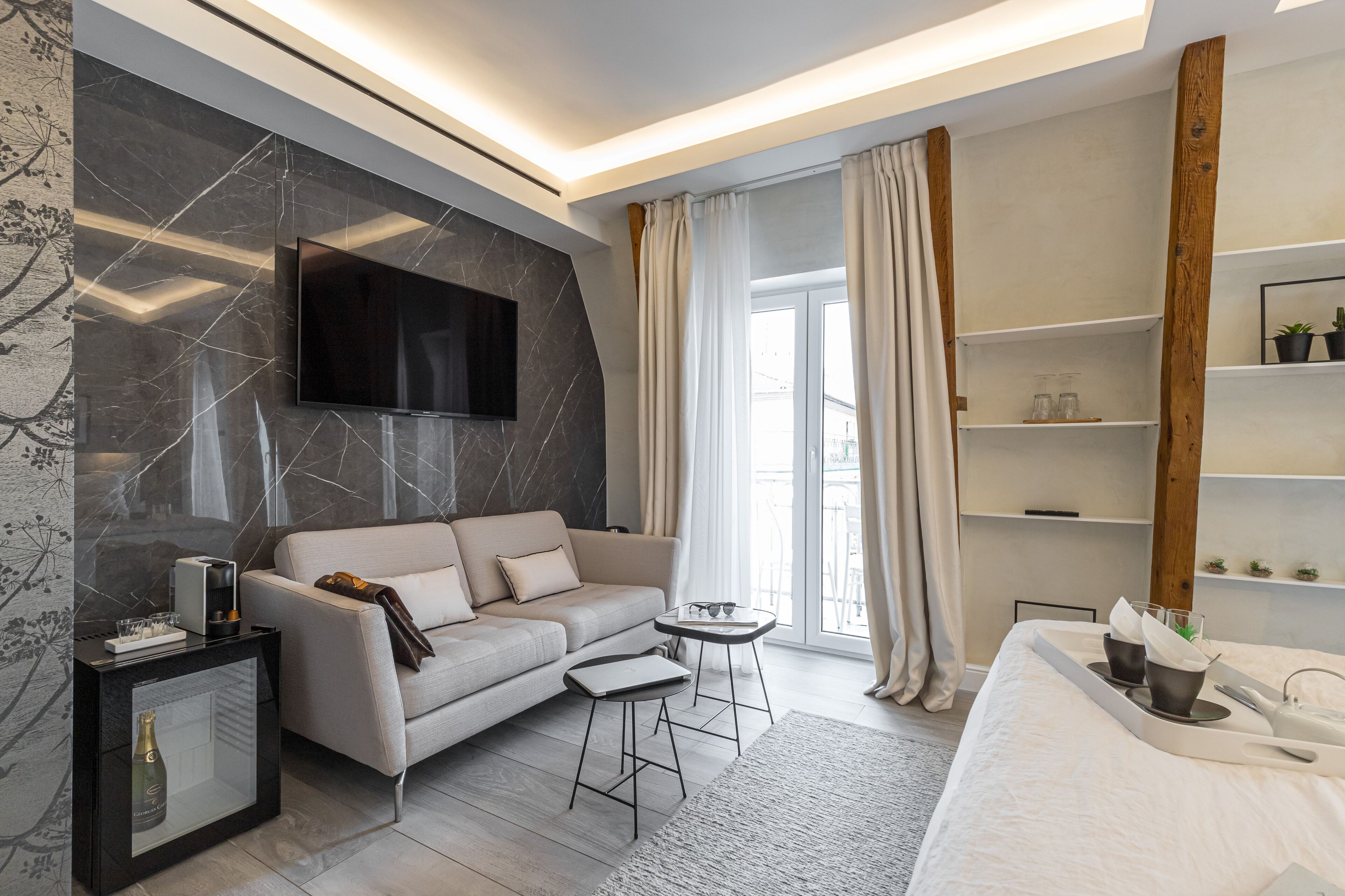 Een moderne hotelkamer met een bank, een klein tafeltje, een televisie bevestigd aan een marmeren muur en een bed met een dienblad met verzorgingsproducten.