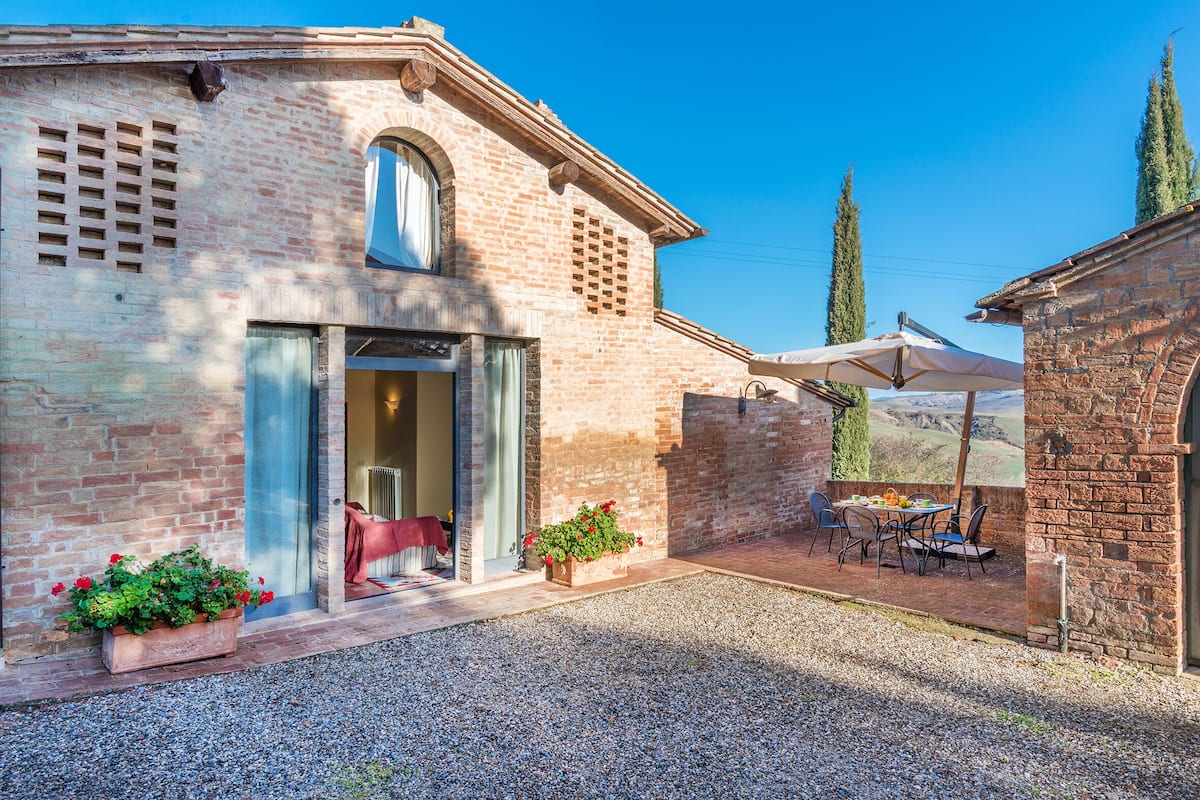 A rustic brick building with a glass door entrance, a patio with outdoor seating, and a view of the countryside.