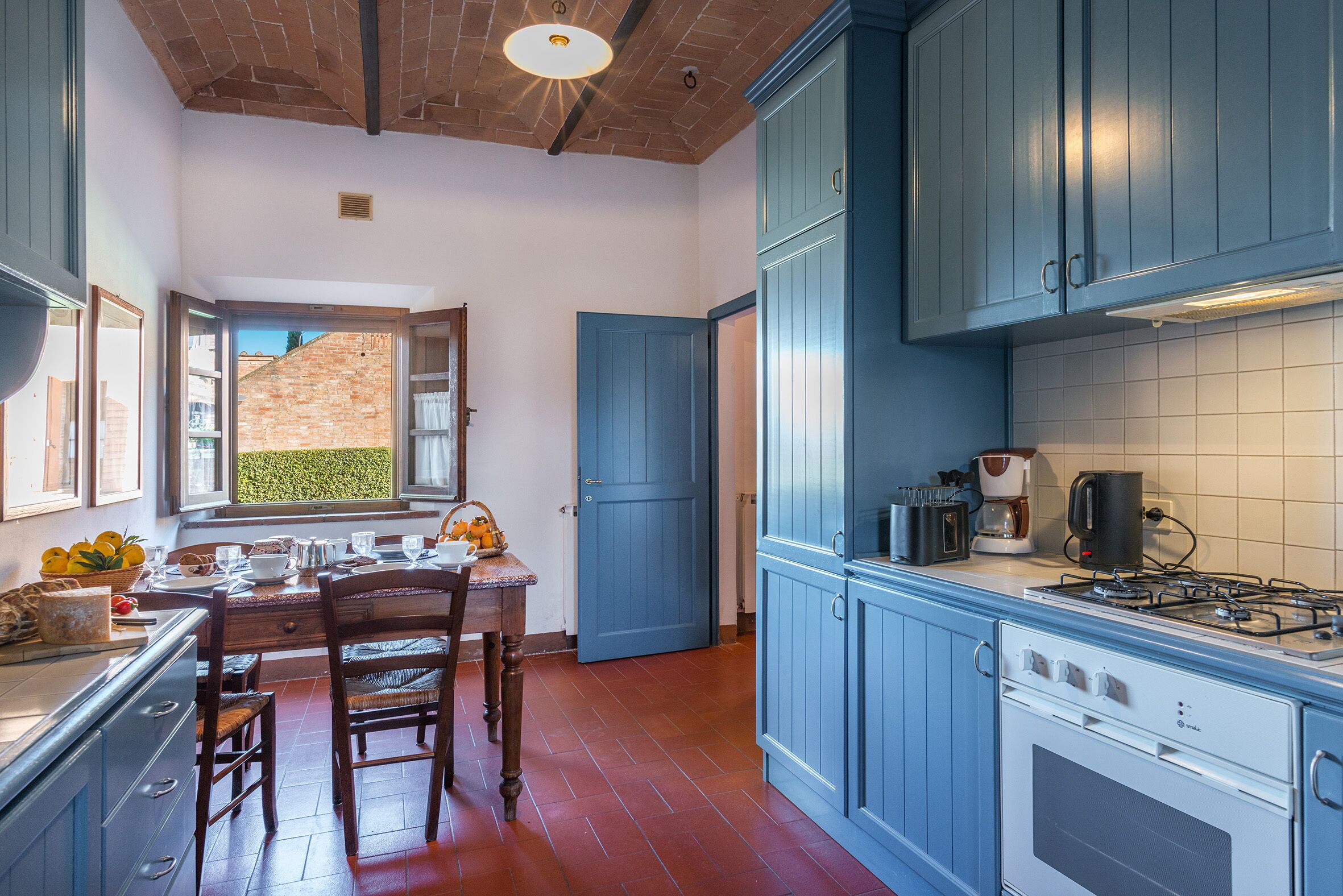 Une cuisine avec des meubles de rangement bleus, une table à manger en bois avec des chaises, et une vue sur un mur de briques à travers une porte ouverte.